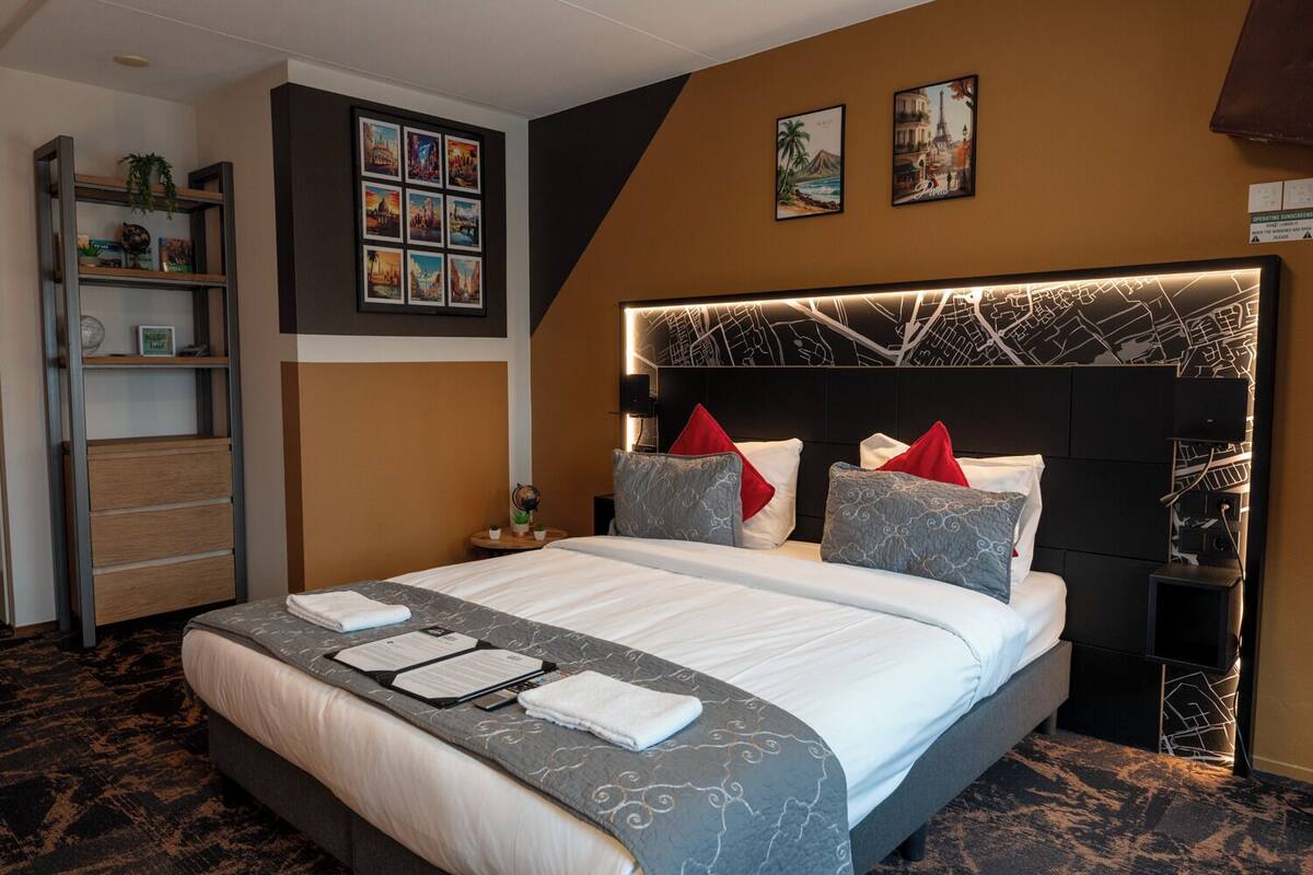 A hotel room with a large bed, a headboard with a map design, a bookshelf, and two framed pictures on the wall.