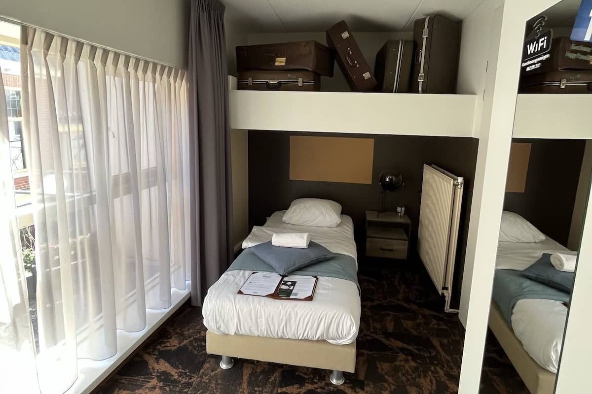 A compact hotel room with a bunk bed, a desk, a radiator, and a window with curtains.