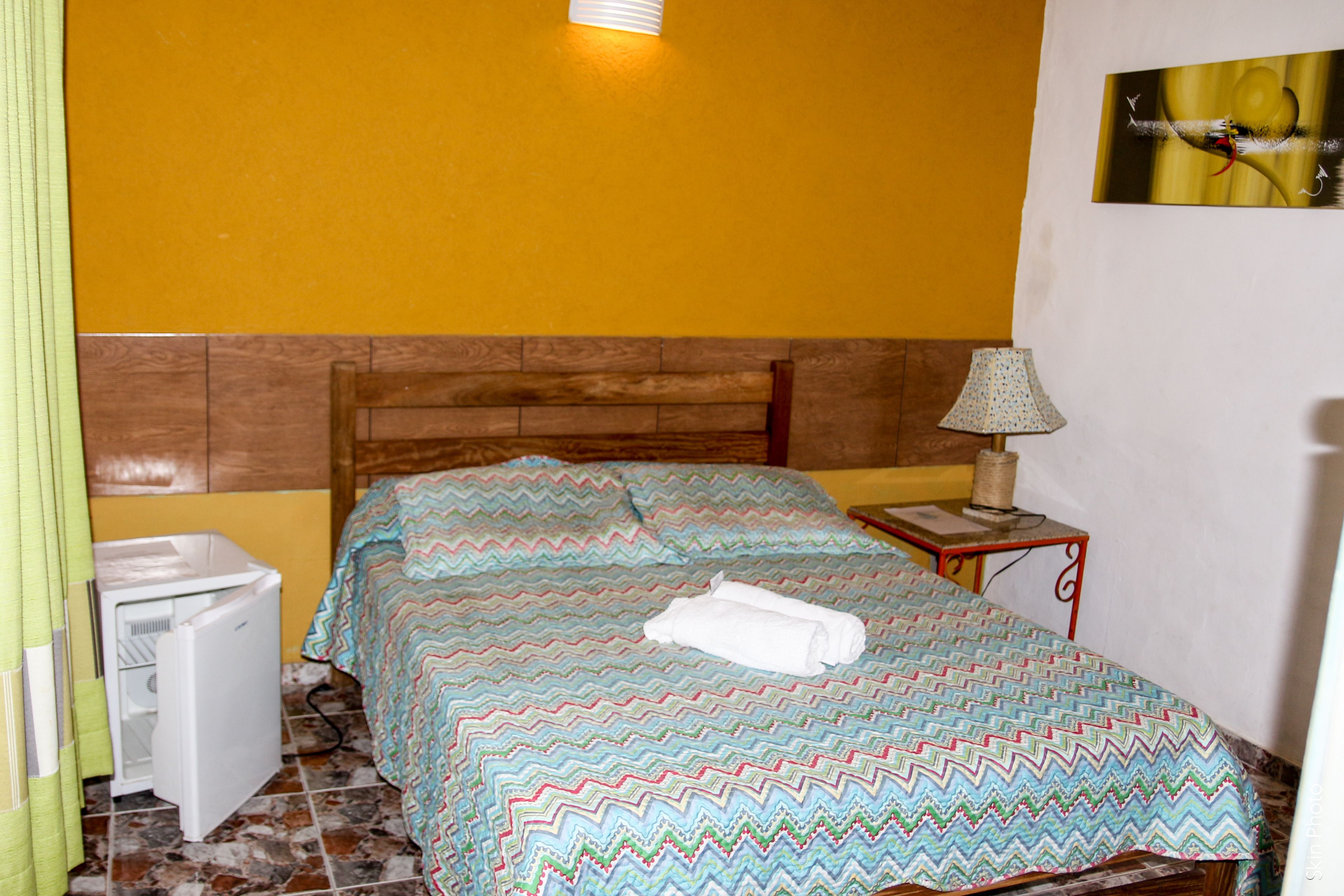 Ein Schlafzimmer mit einem Bett, einem Nachttisch mit Lampe, einem kleinen Kühlschrank und einem an der Wand hängenden Bild.