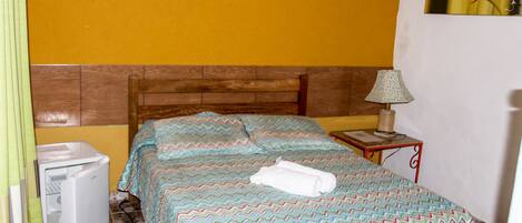 A bedroom with a bed, a bedside table with a lamp, a small refrigerator, and a wall-mounted picture.