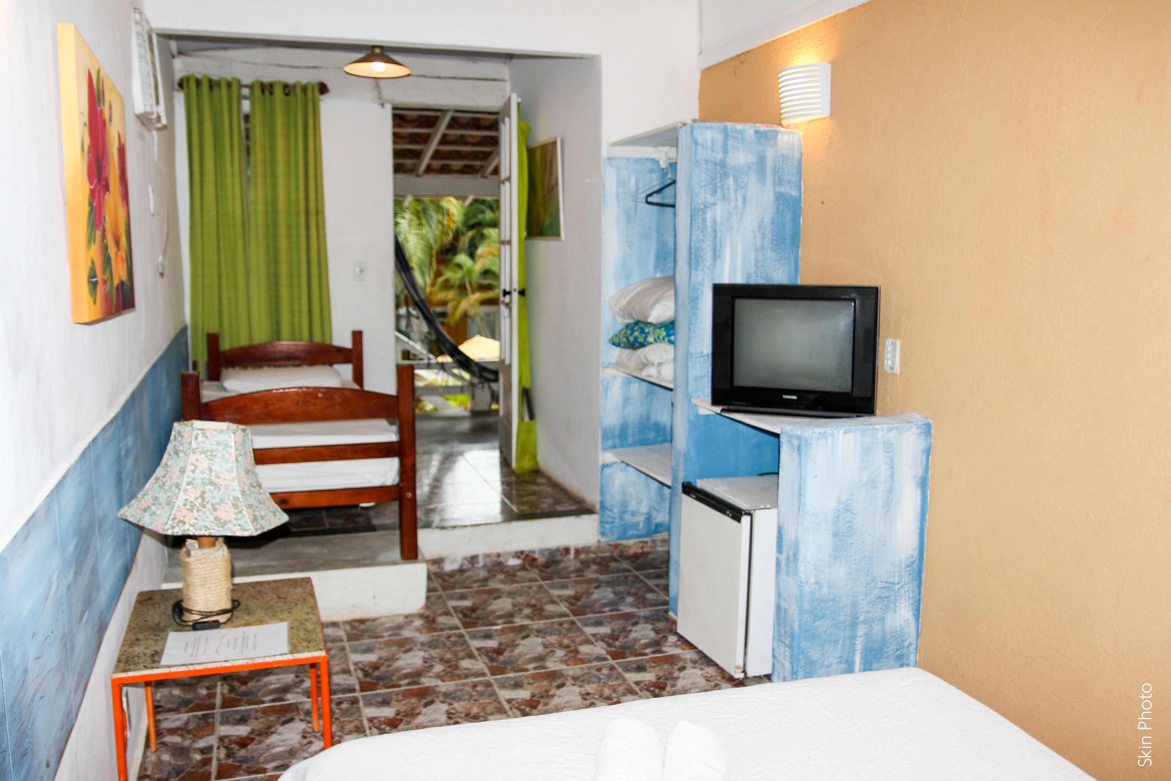 Una camera compatta con un letto, un piccolo tavolo con una lampada, una televisione su un mobile e una porta che dà su un balcone con vista su palme.