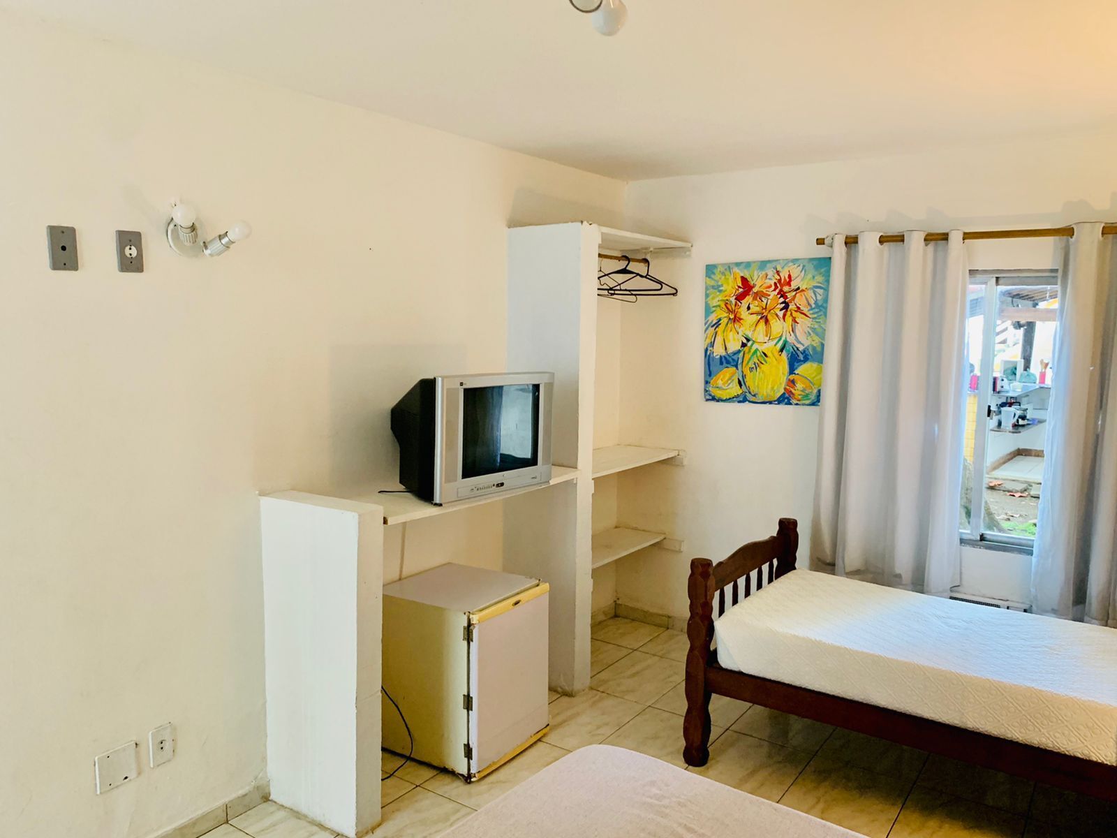 Habitación compacta con cama, un pequeño televisor en un estante, un refrigerador y un cuadro en la pared.