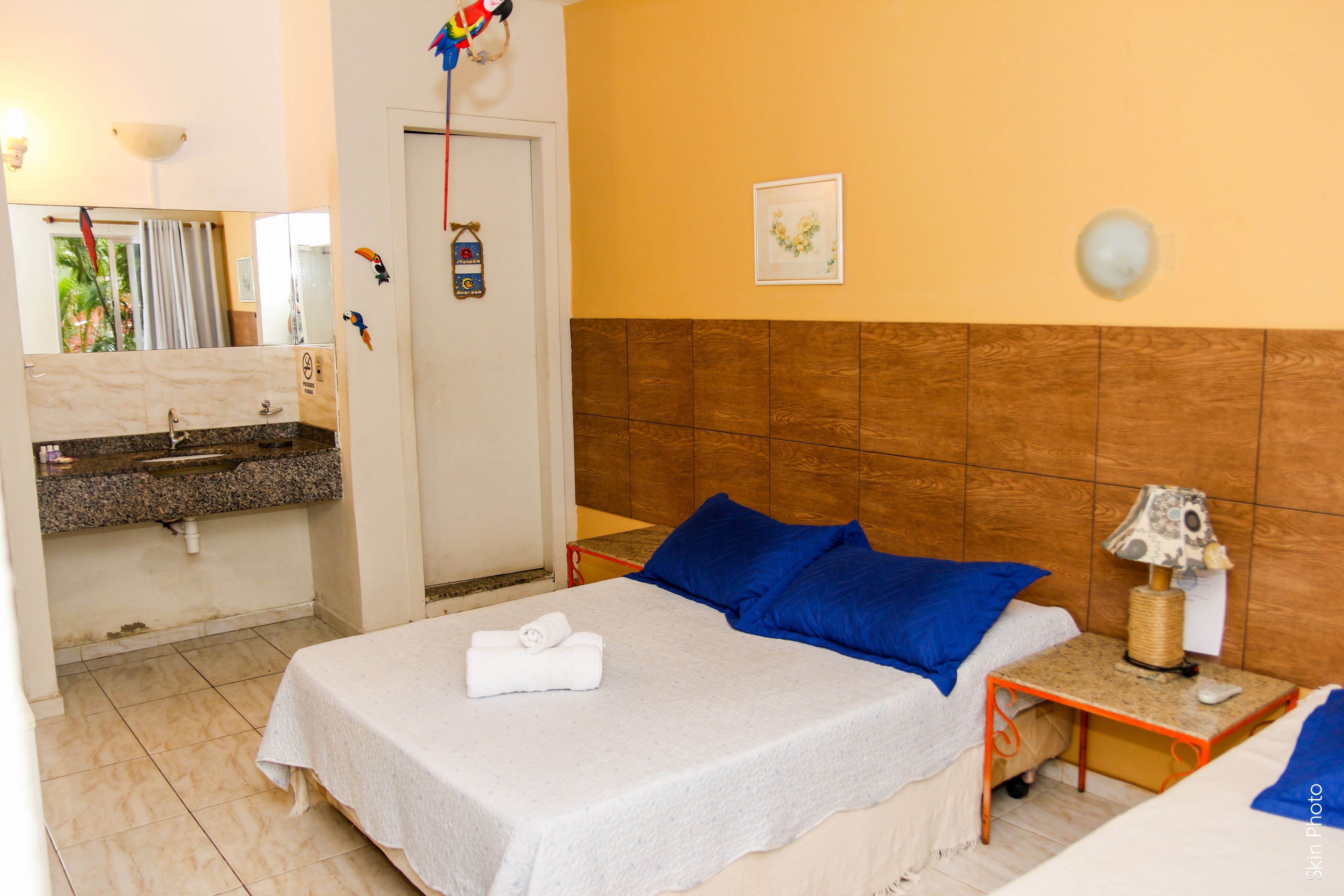 A hotel room with a bed, a nightstand, a lamp, a wall clock, a mirror, and a bathroom with a sink and towel.