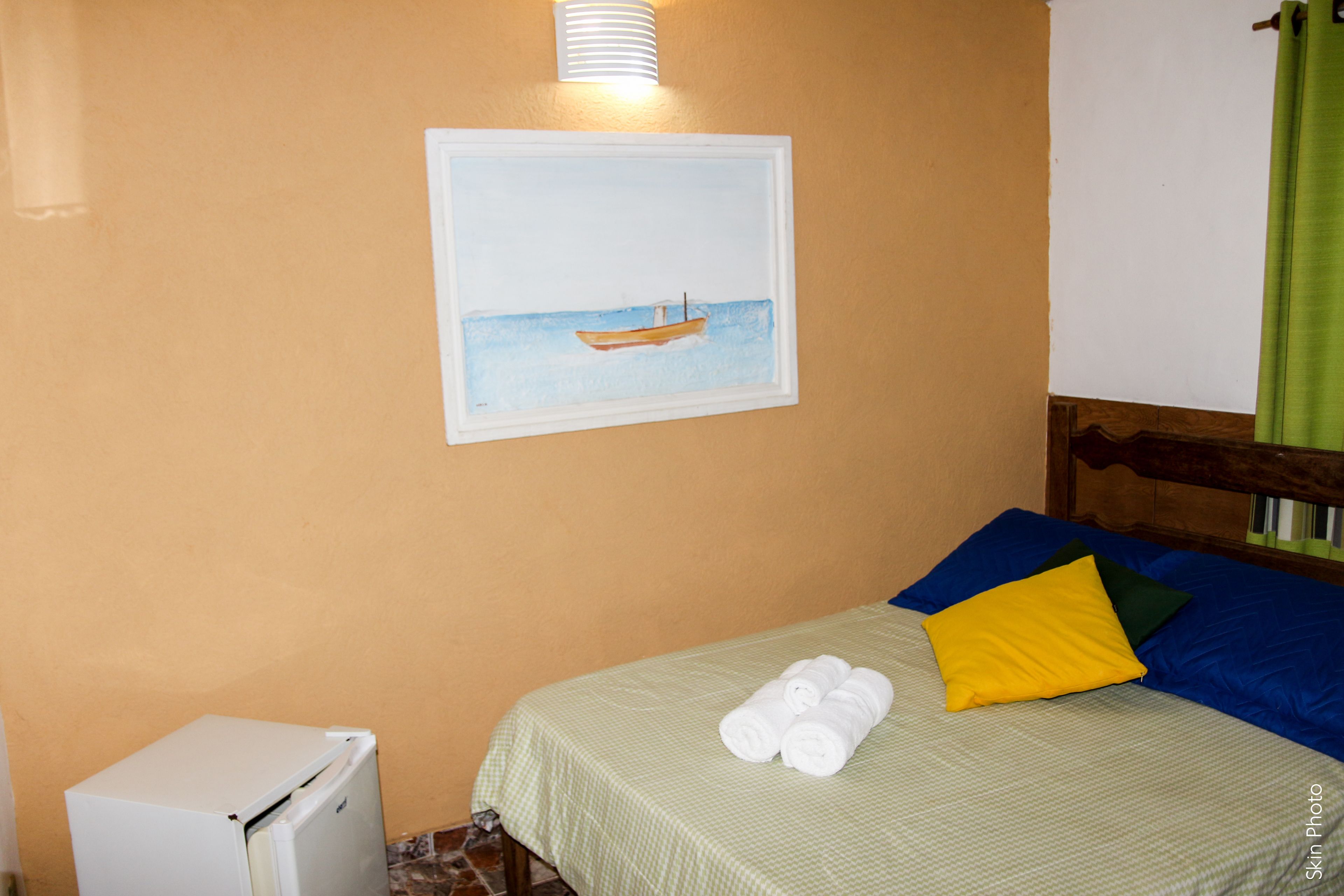 A bedroom with a bed, pillows, a small white refrigerator, and a painting of a boat on the wall.