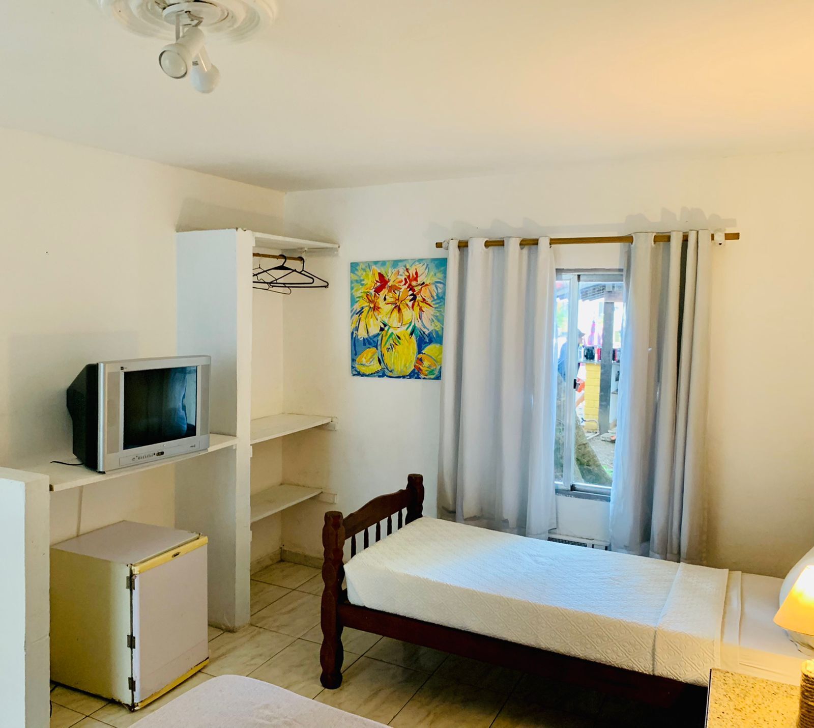 Quarto pequeno e bem iluminado, com uma cama, televisão, guarda-roupa e janela com cortinas.