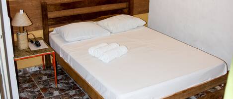 Una cama individual con ropa de cama blanca, cabecera de madera, mesita de noche con lámpara y ventilador de techo.