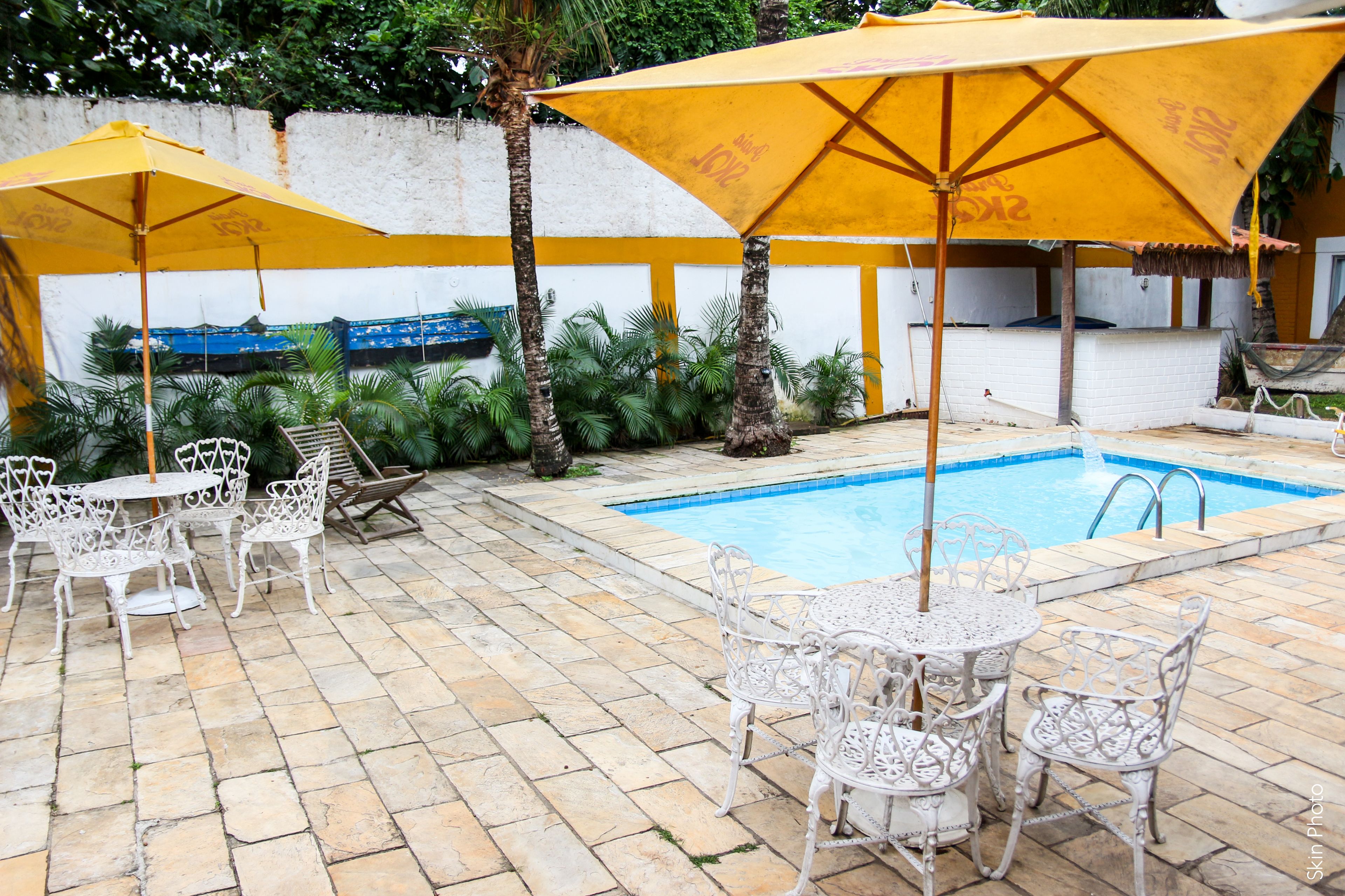 Zona de piscina con sillas y mesas de metal blanco, sombrillas amarillas y una piscina.