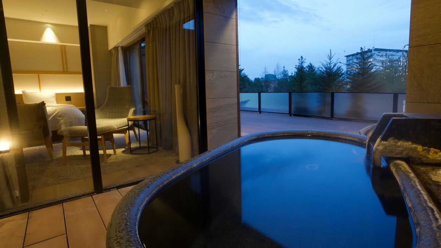 A hotel room with a glass partition, a bed, a chair, and a view of a balcony with a pool.