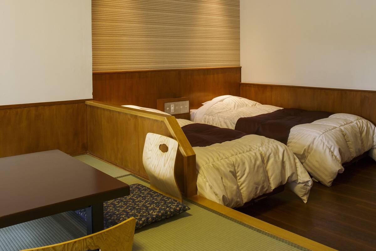 A room with a wooden ceiling, a bed with pillows, a table with a chair, and a tatami mat.