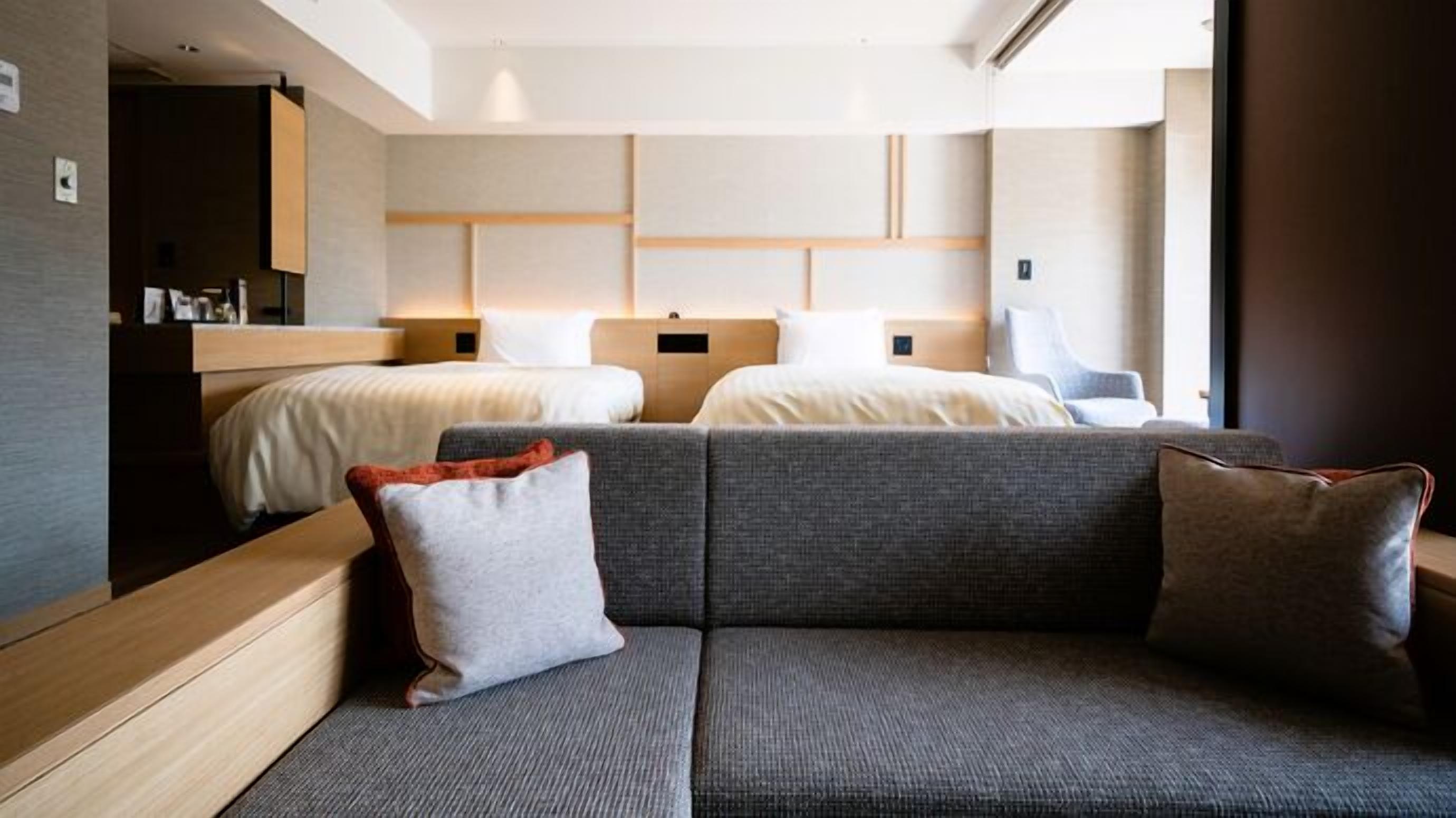 A modern hotel room with a sofa, two beds, a wooden headboard, and a small desk.