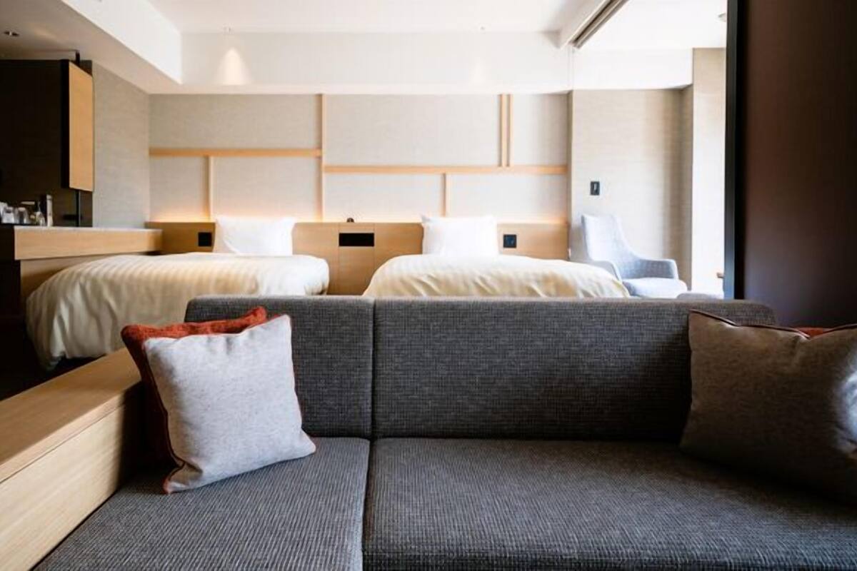 A modern hotel room with a sofa, two beds, a wooden headboard, and a small desk.
