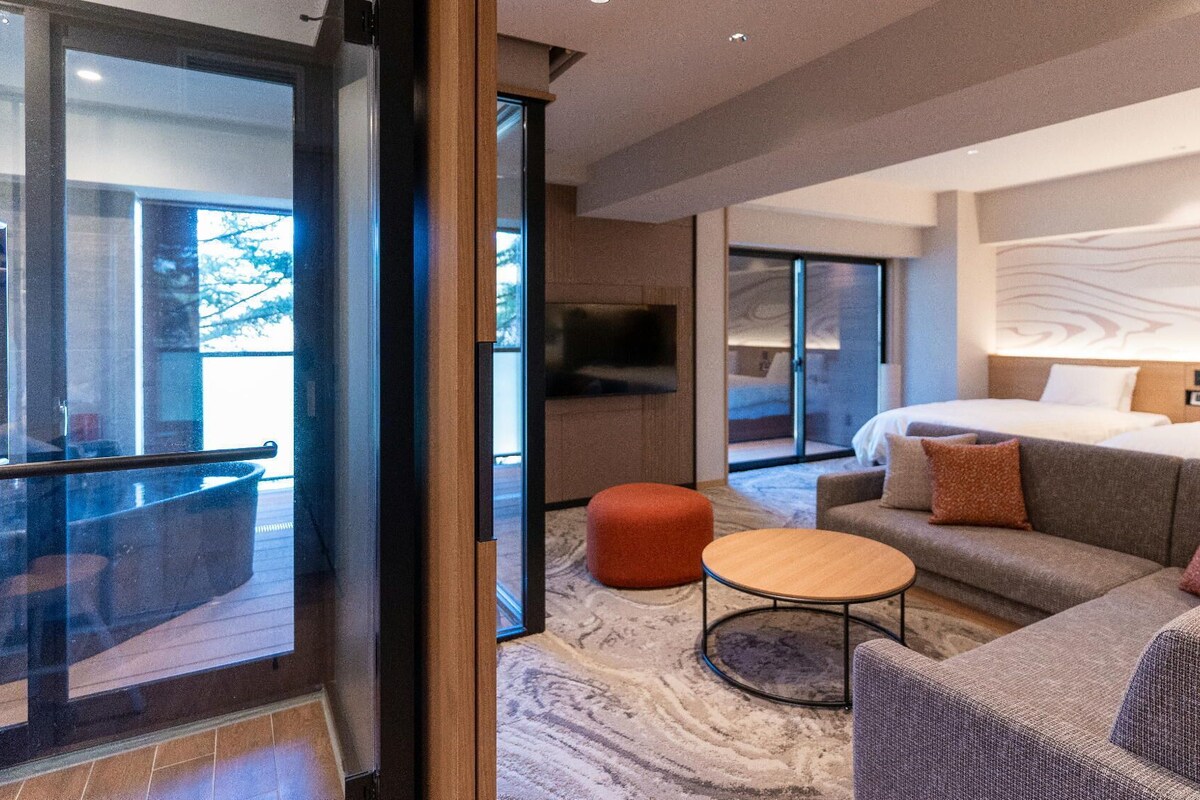 Une chambre d’hôtel moderne dotée d’un grand lit, d’un canapé, d’une table basse ronde et d’une porte vitrée donnant sur un balcon.
