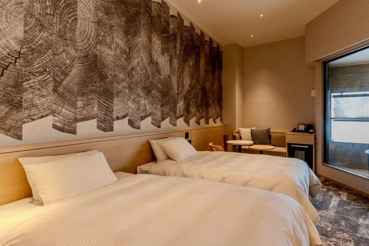 A hotel room with two beds, a small table, a chair, and a large window.
