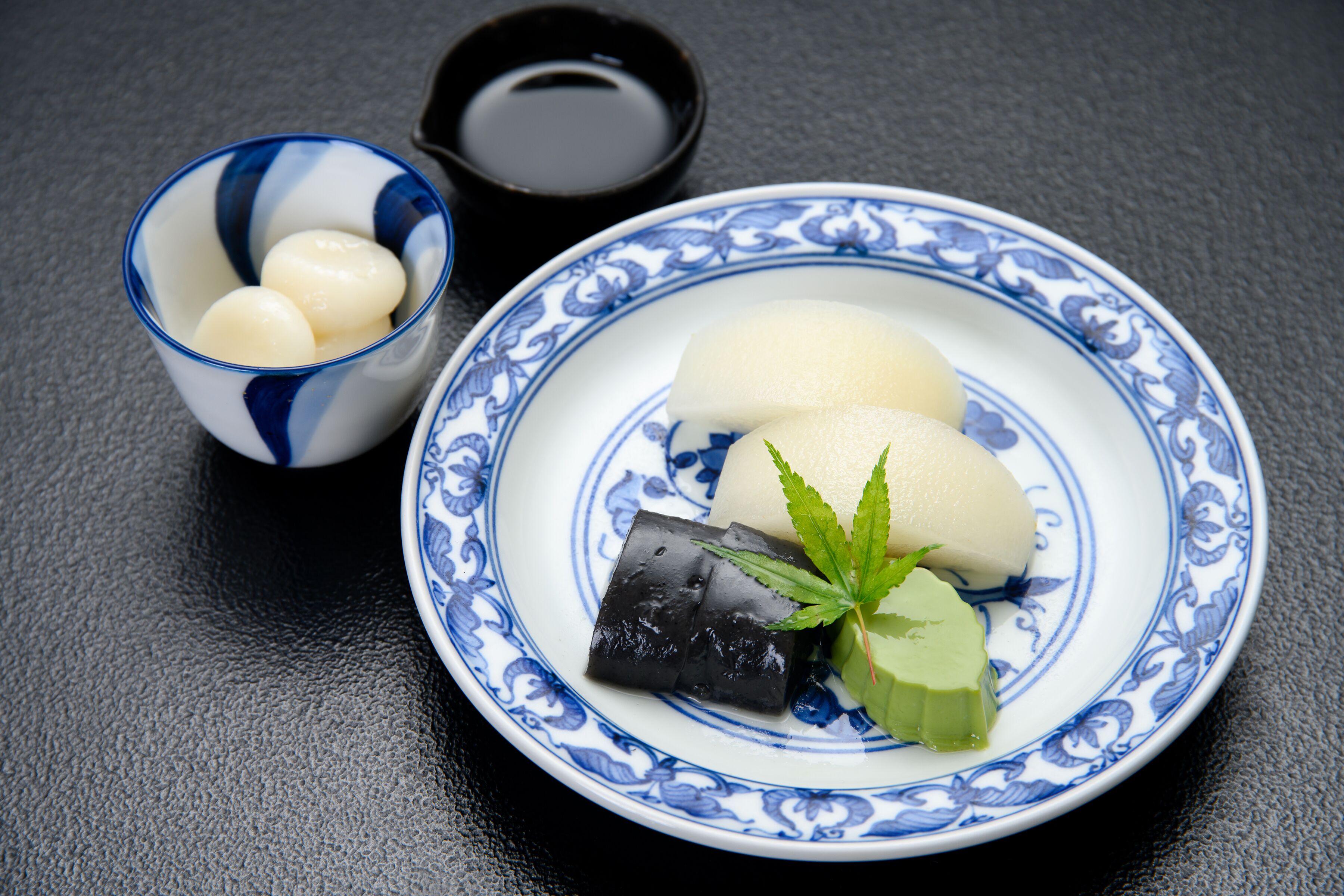 A plate with three white dumplings and a green vegetable, accompanied by a small bowl of white dumplings and a cup of soy sauce.