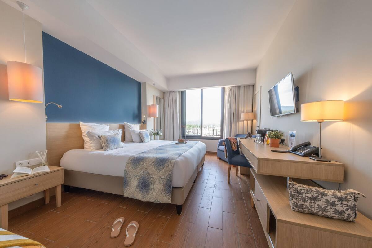 A hotel room with a large bed, a desk, a chair, a TV, and a window with a view.