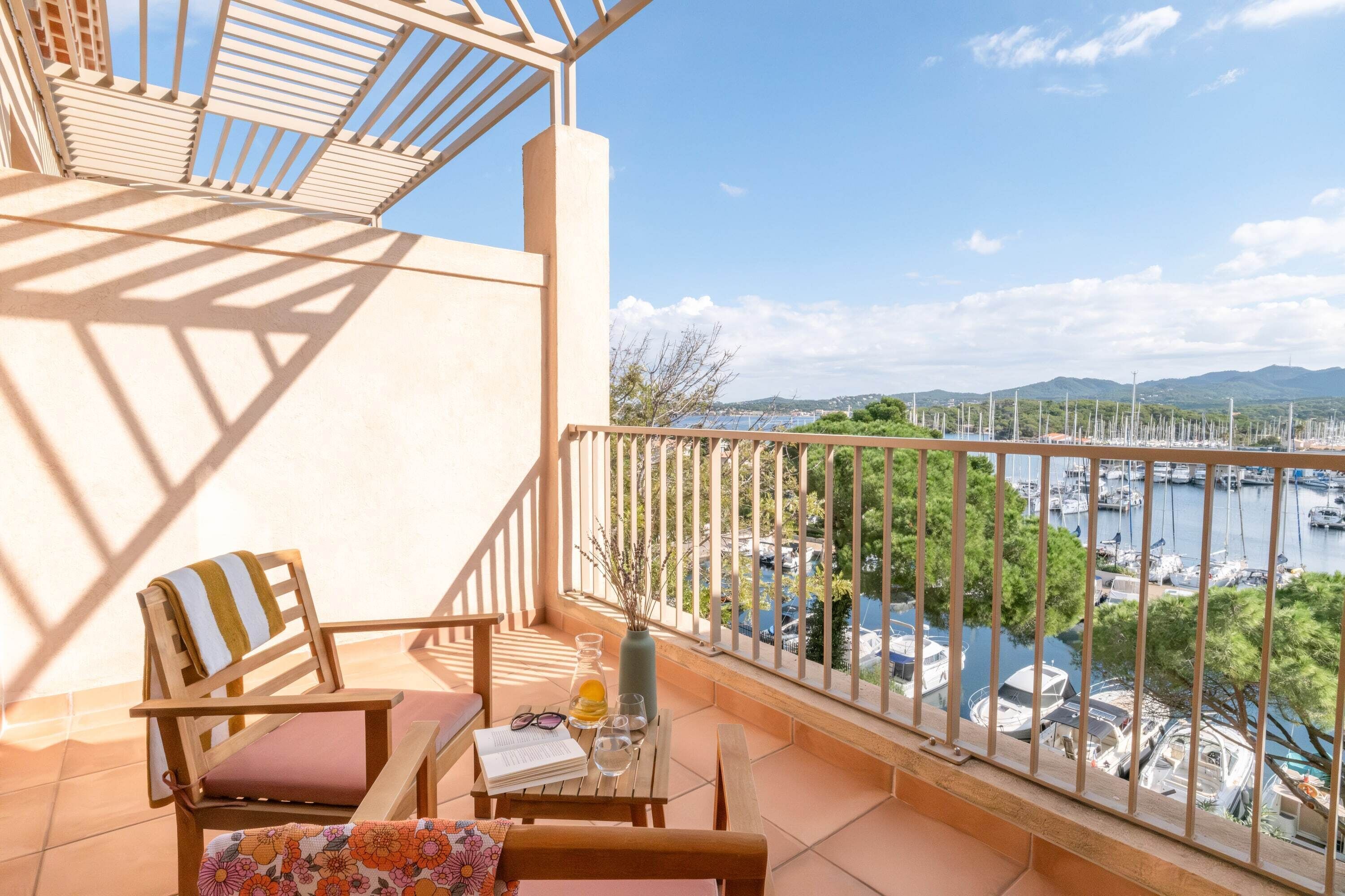 A balcony with a wooden chair, a small table with a book, a glass of water, and a vase with branches. The balcony overlooks a marina with boats and greenery.