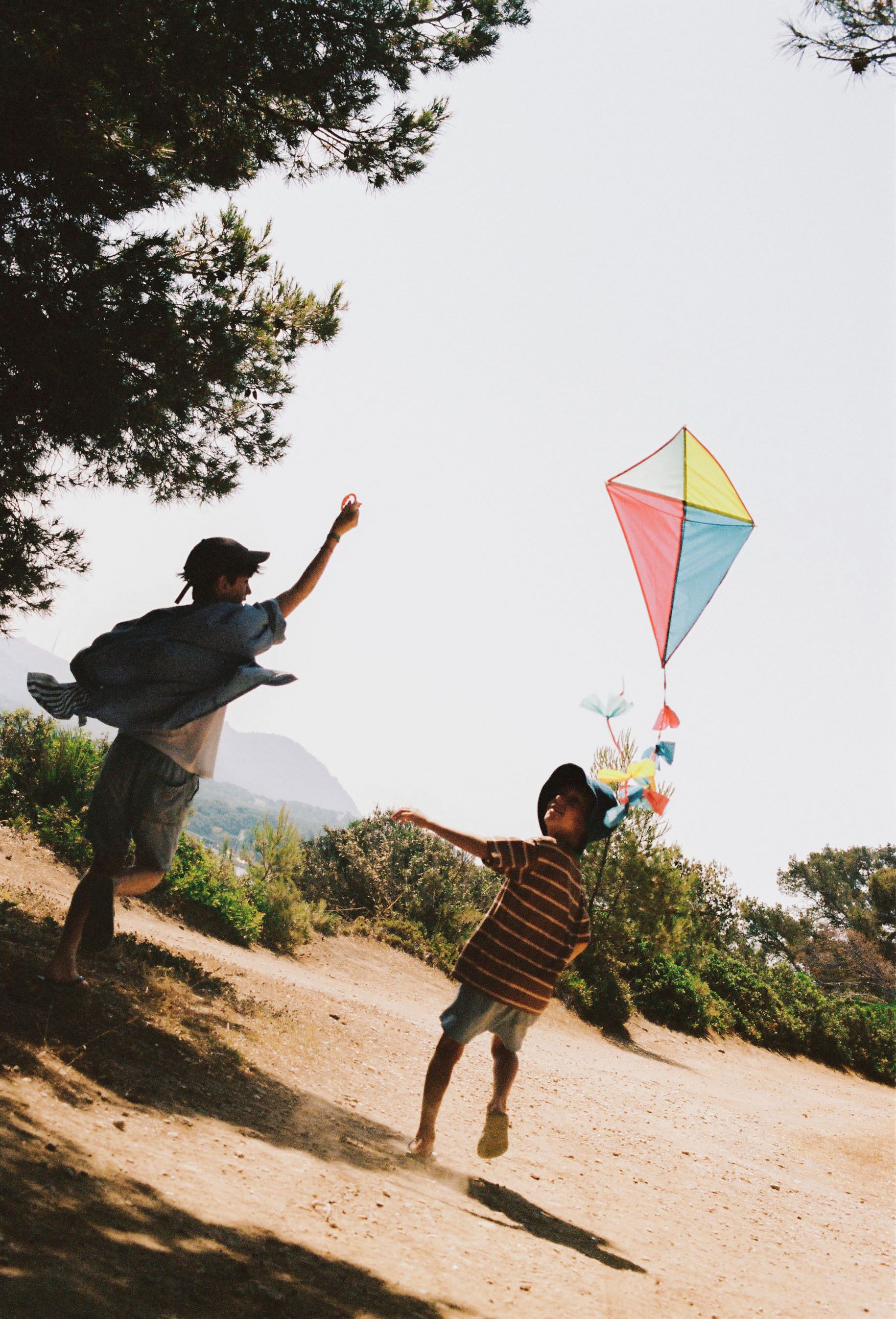 Two people flying a kite on a dirt path with trees and mountains in the background.