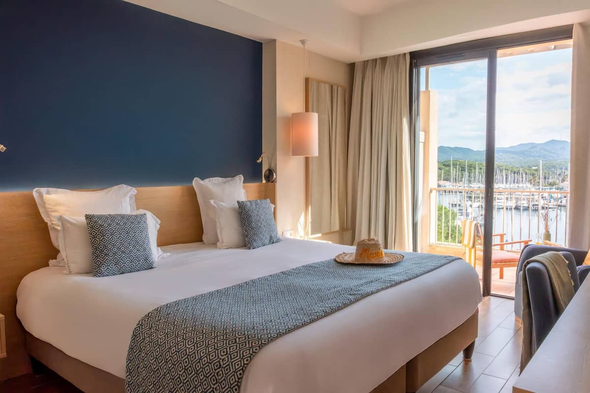 A hotel room with a large bed, a view of a marina, and a balcony with a chair.