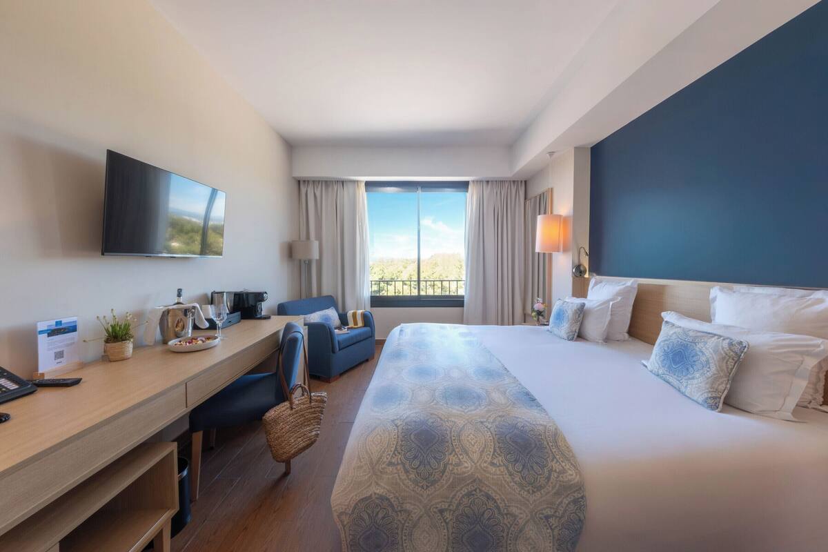 A hotel room with a large bed, a desk with a computer and a lamp, a TV mounted on the wall, and a view of the outdoors through the window.