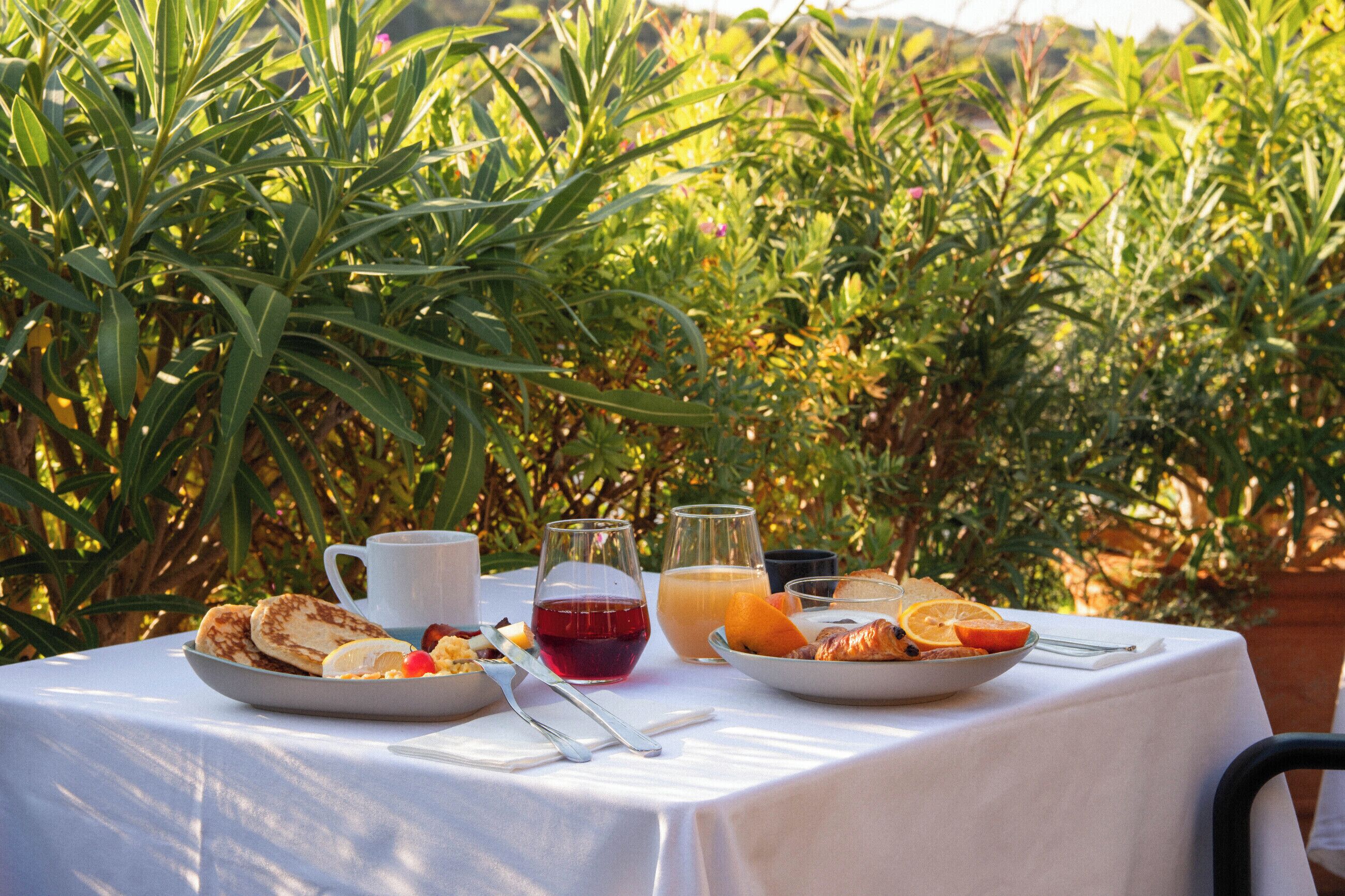 Mesa al aire libre con desayuno, incluyendo panqueques, rodajas de naranja y jugo, rodeada de follaje.