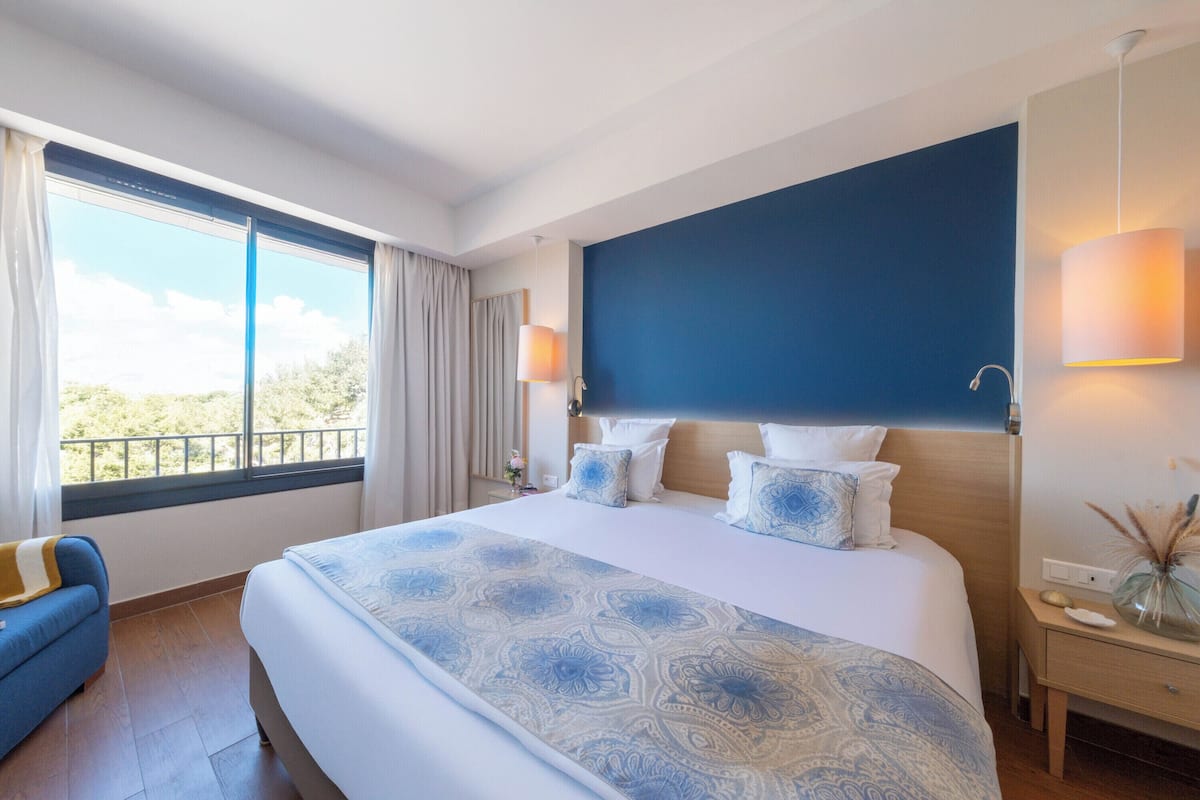 A hotel room with a large bed, a blue accent wall, a window with curtains, a bedside table with a lamp, and a view of greenery outside.