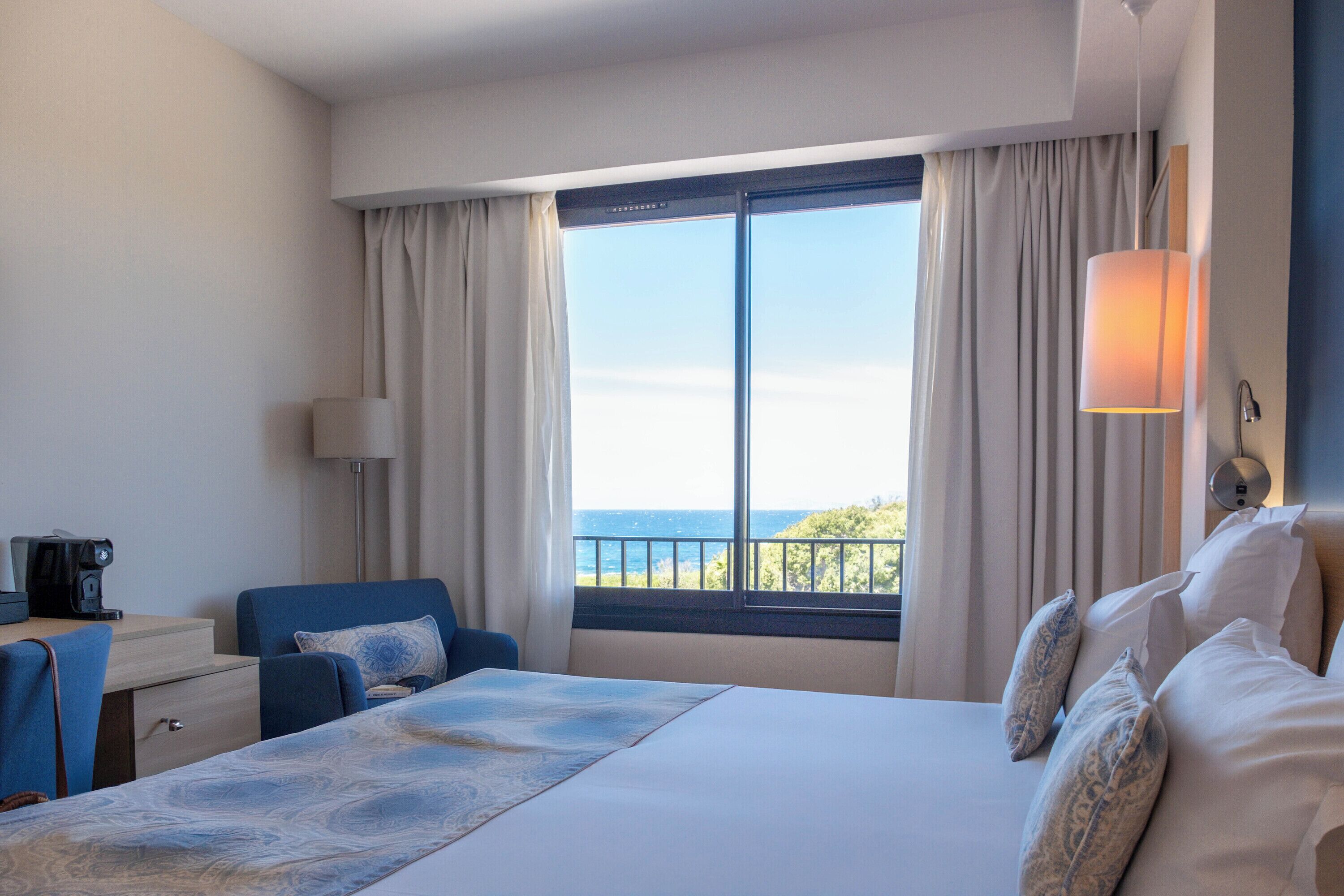 A hotel room with a bed, a desk, a chair, a lamp, and a view of the sea.