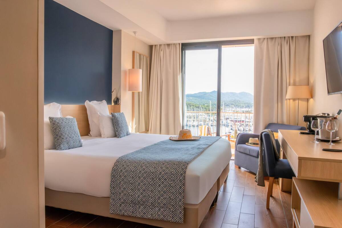 A hotel room with a large bed, a desk, a chair, and a view of the city through the window.