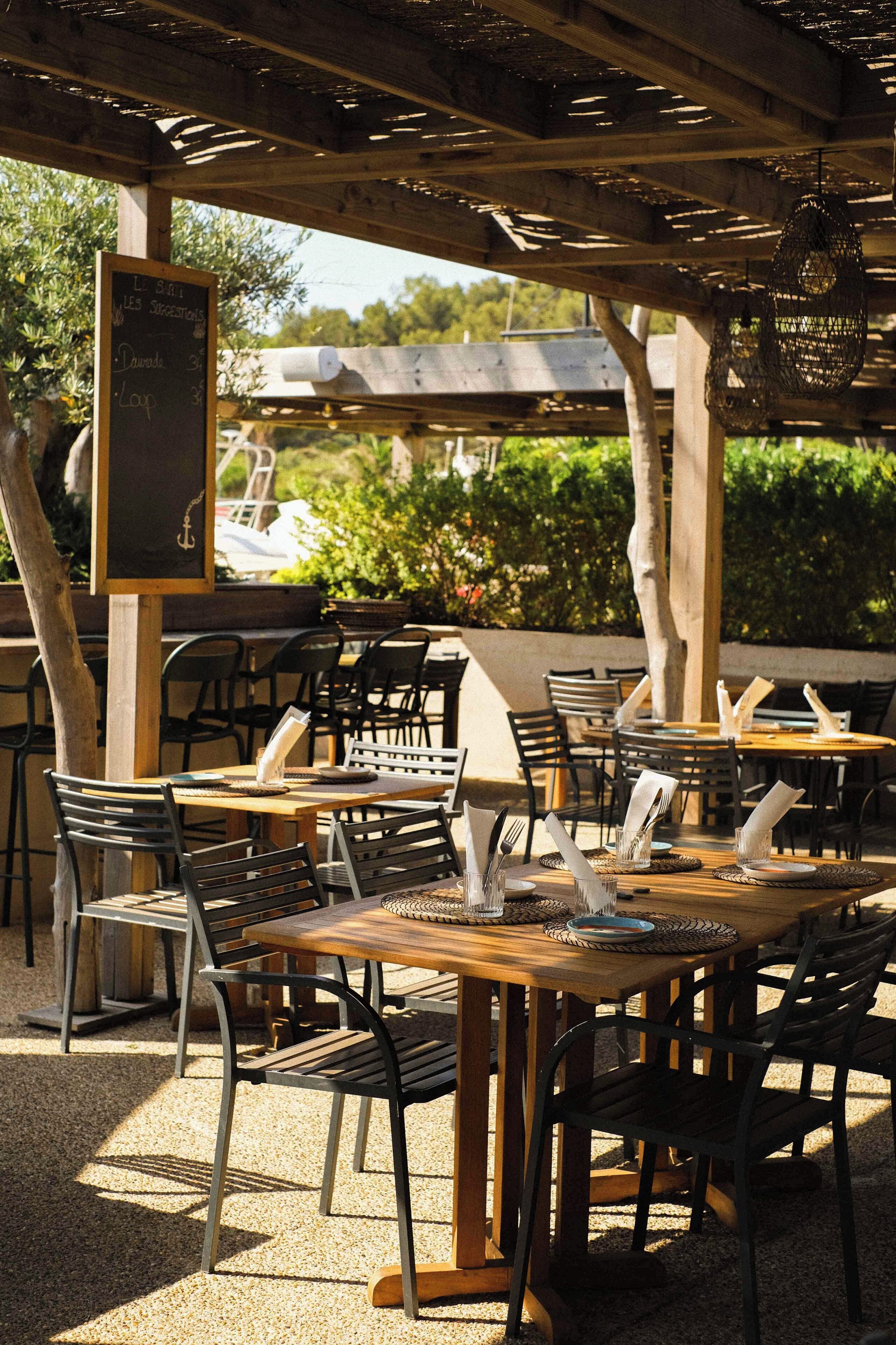 An outdoor cafe with wooden tables and metal chairs, a chalkboard menu, and a pergola overhead.