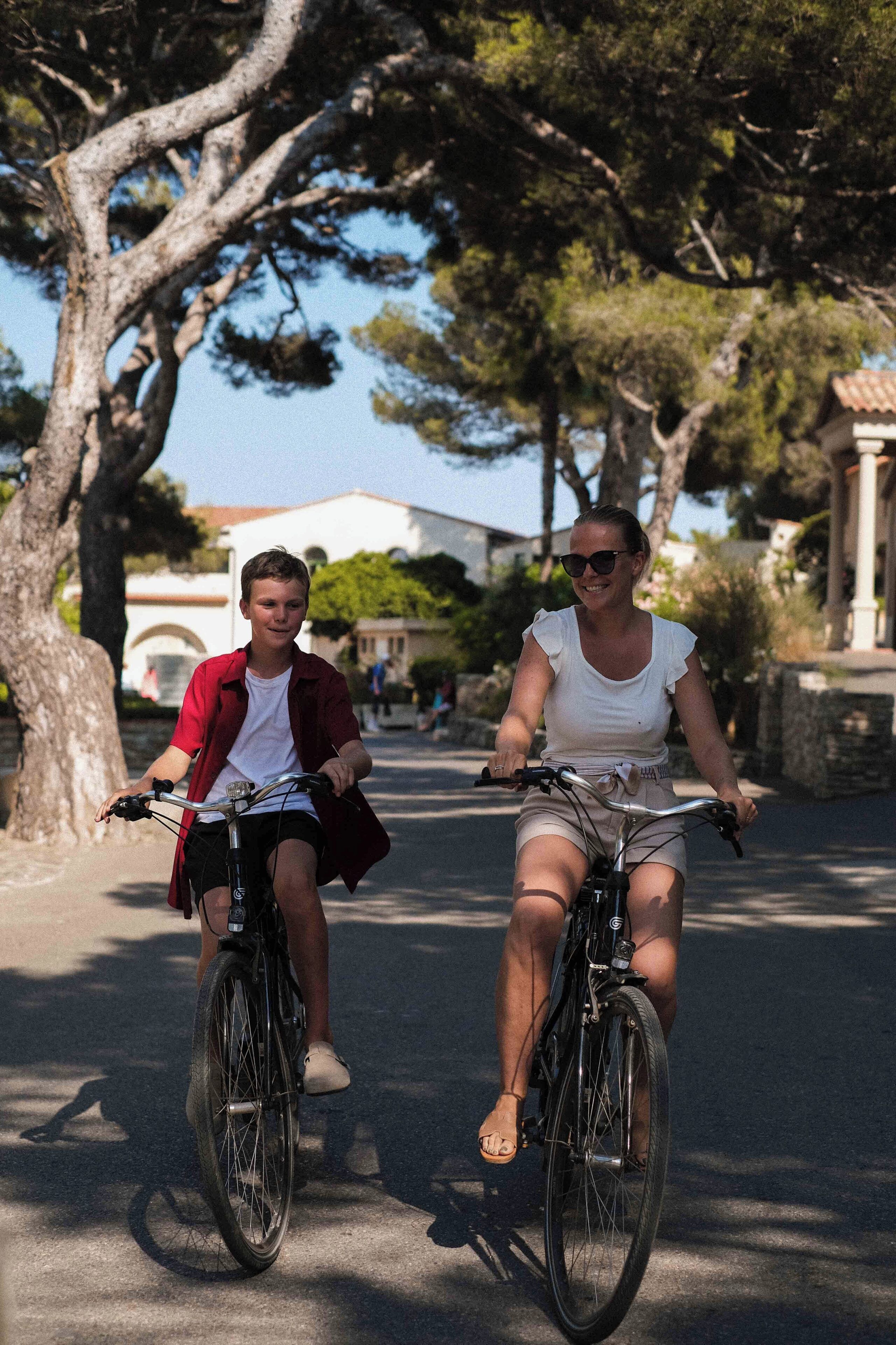 Two people riding bicycles on a street with trees and buildings in the background.