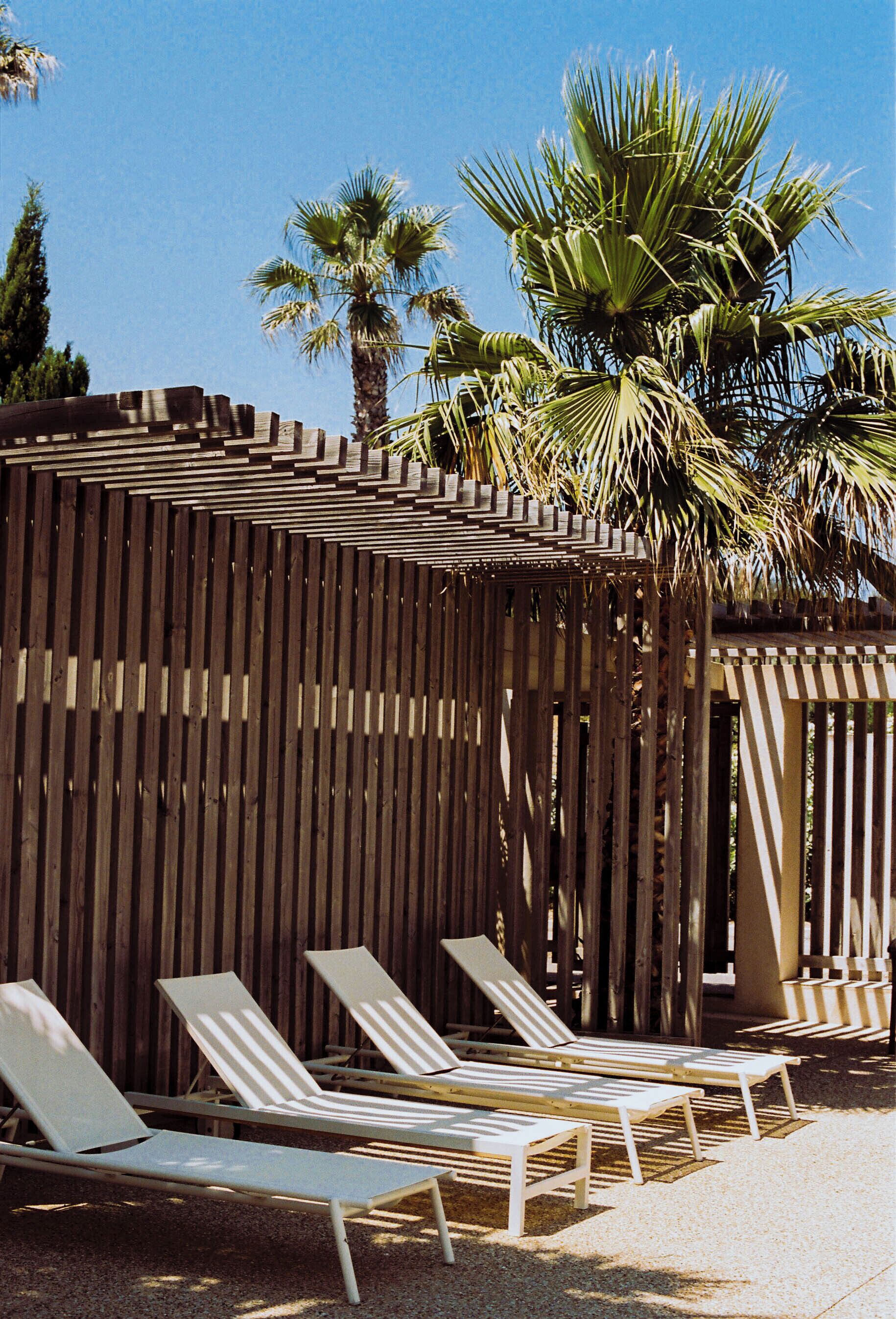 Une clôture en bois, avec un palmier derrière et trois chaises longues blanches devant.