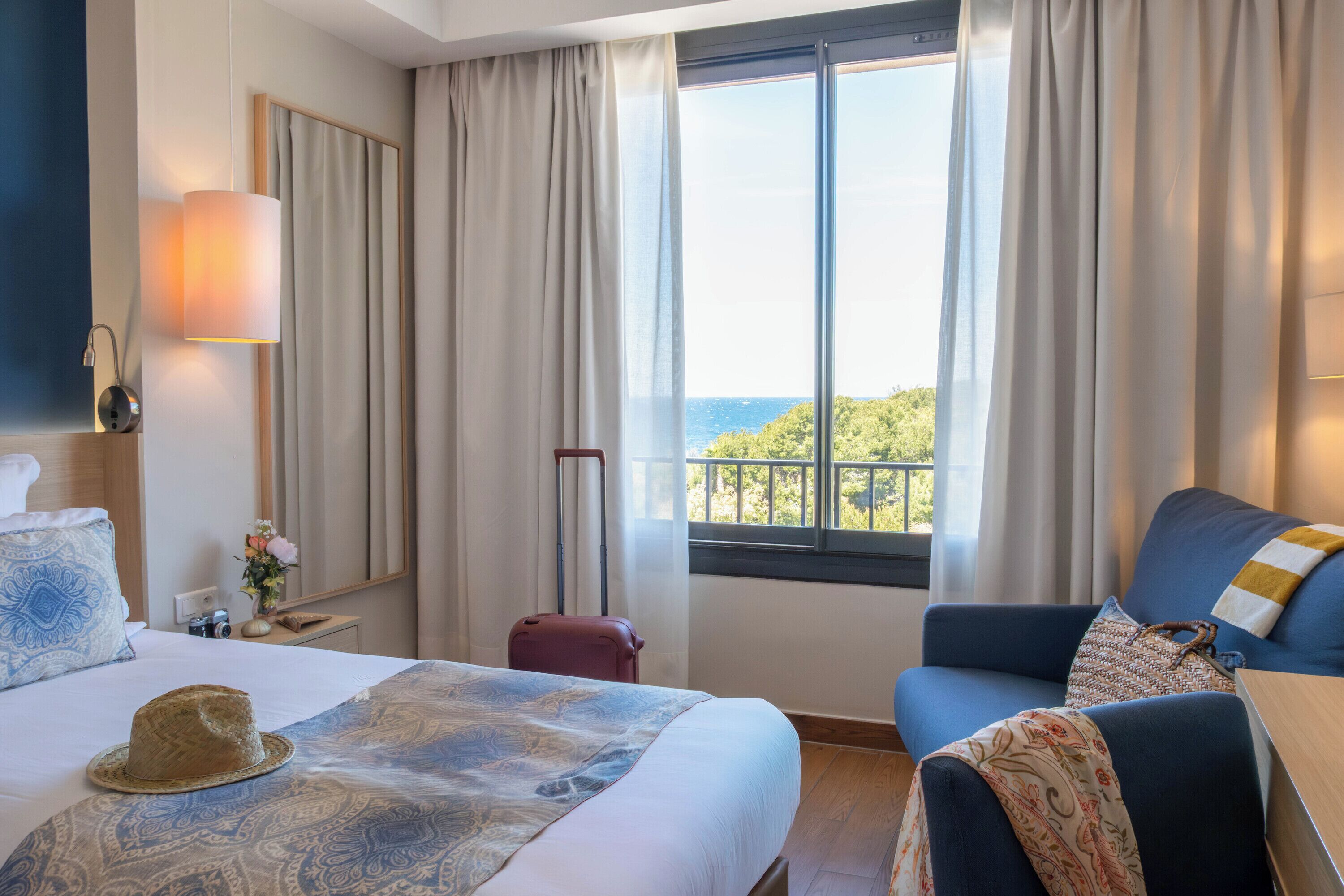 A hotel room with a bed, a blue armchair, a view of the sea, and a window with curtains.