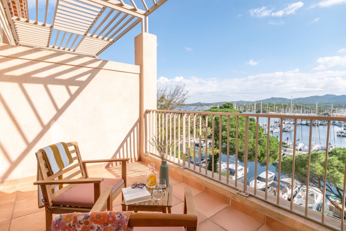 A balcony with a wooden chair, a small table with a book, a glass of water, and a vase with branches. The balcony overlooks a marina with boats and greenery.