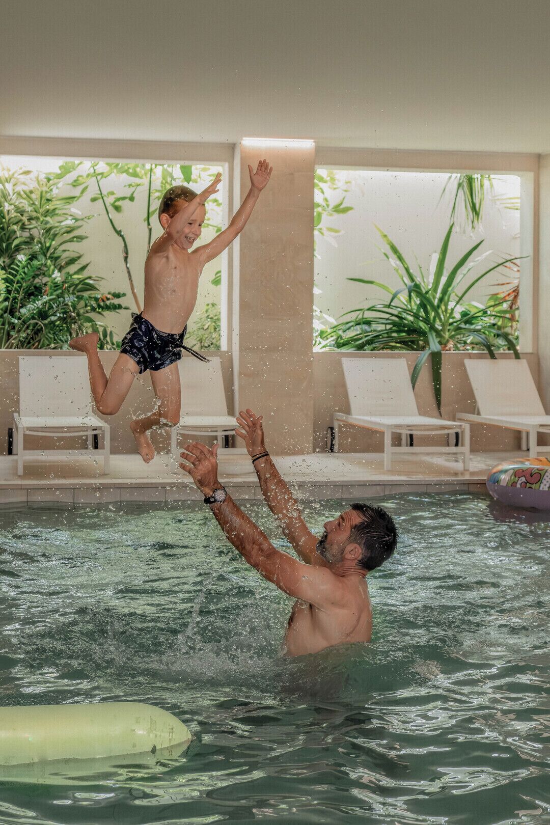 An indoor swimming pool with a child jumping into the water and an adult reaching up to catch them.