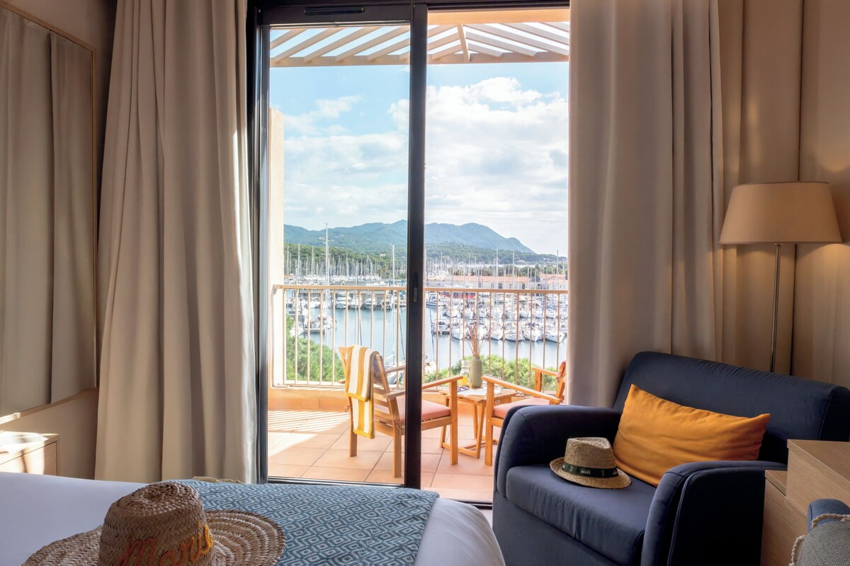 A hotel room with a balcony, a bed, a sofa, a table, and a view of a marina with boats.