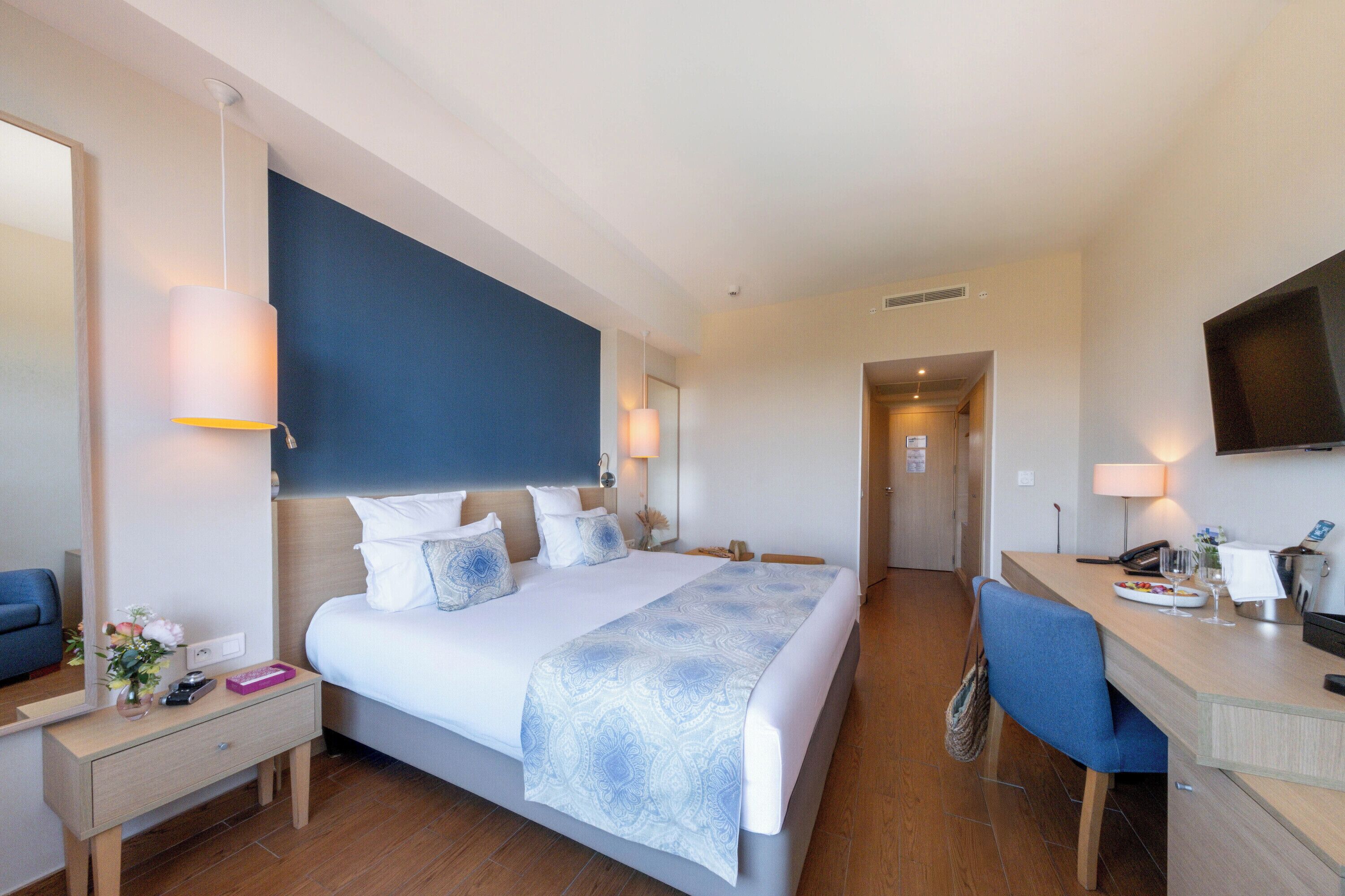 Habitación de hotel con una cama grande, un escritorio, una silla, televisión y un baño visible al fondo.