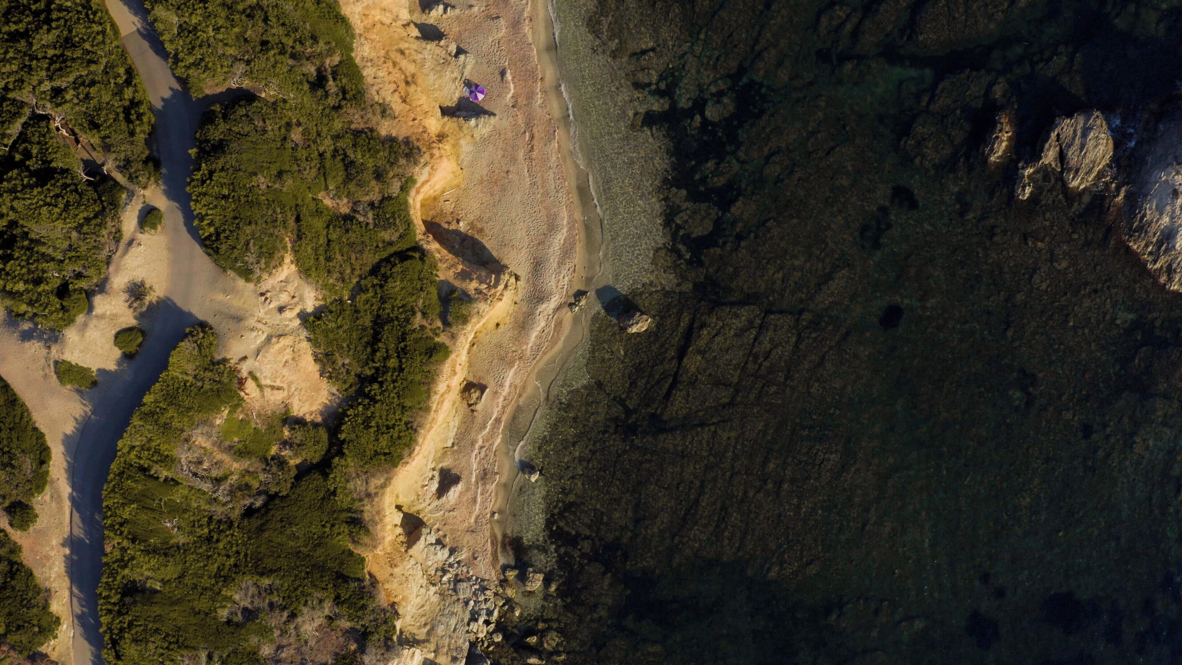 An aerial view of a coastal area with a road, dense greenery, and rocky terrain.