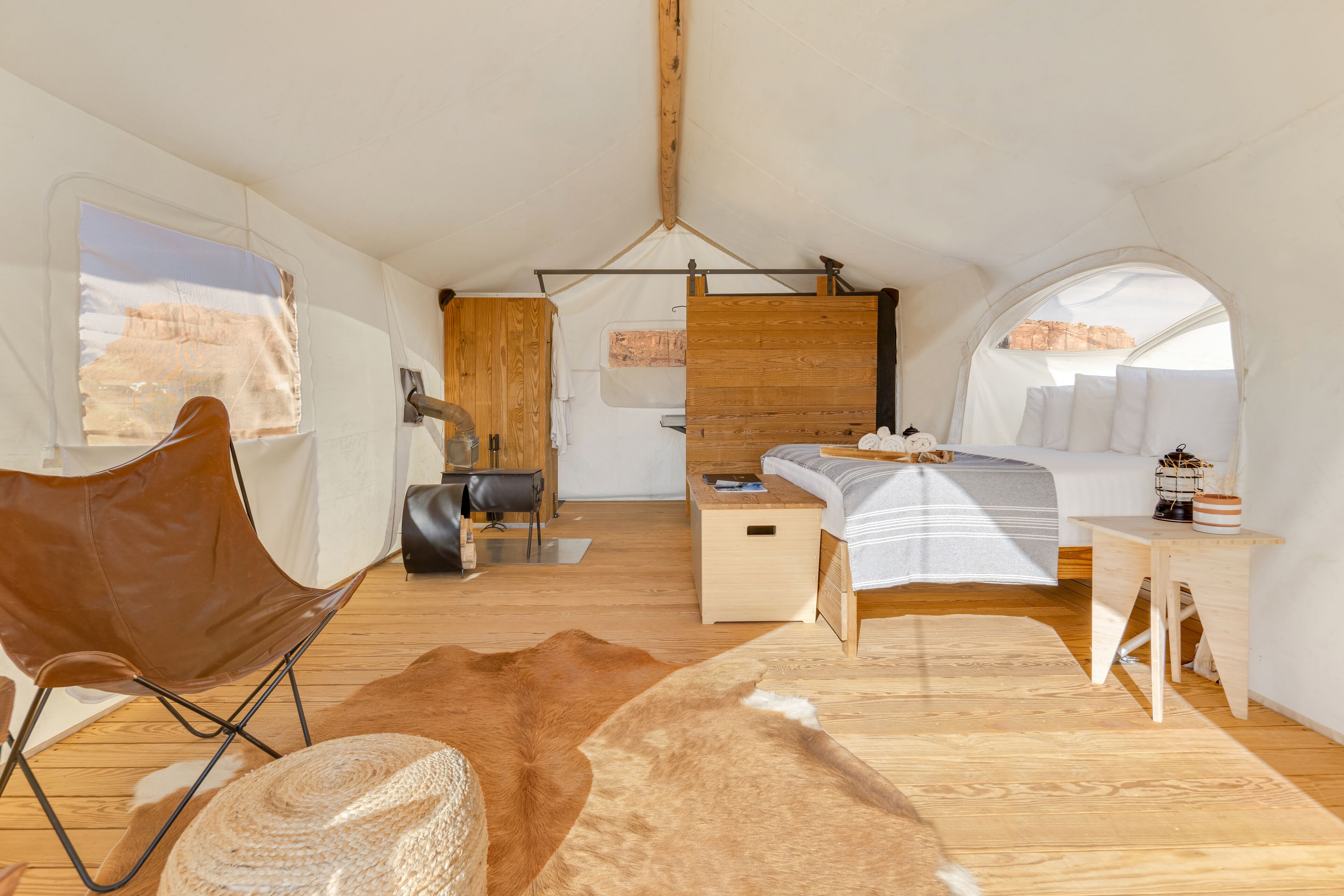 A cozy tented room with a bed, a chair, a small table, and a view of the desert through a window.