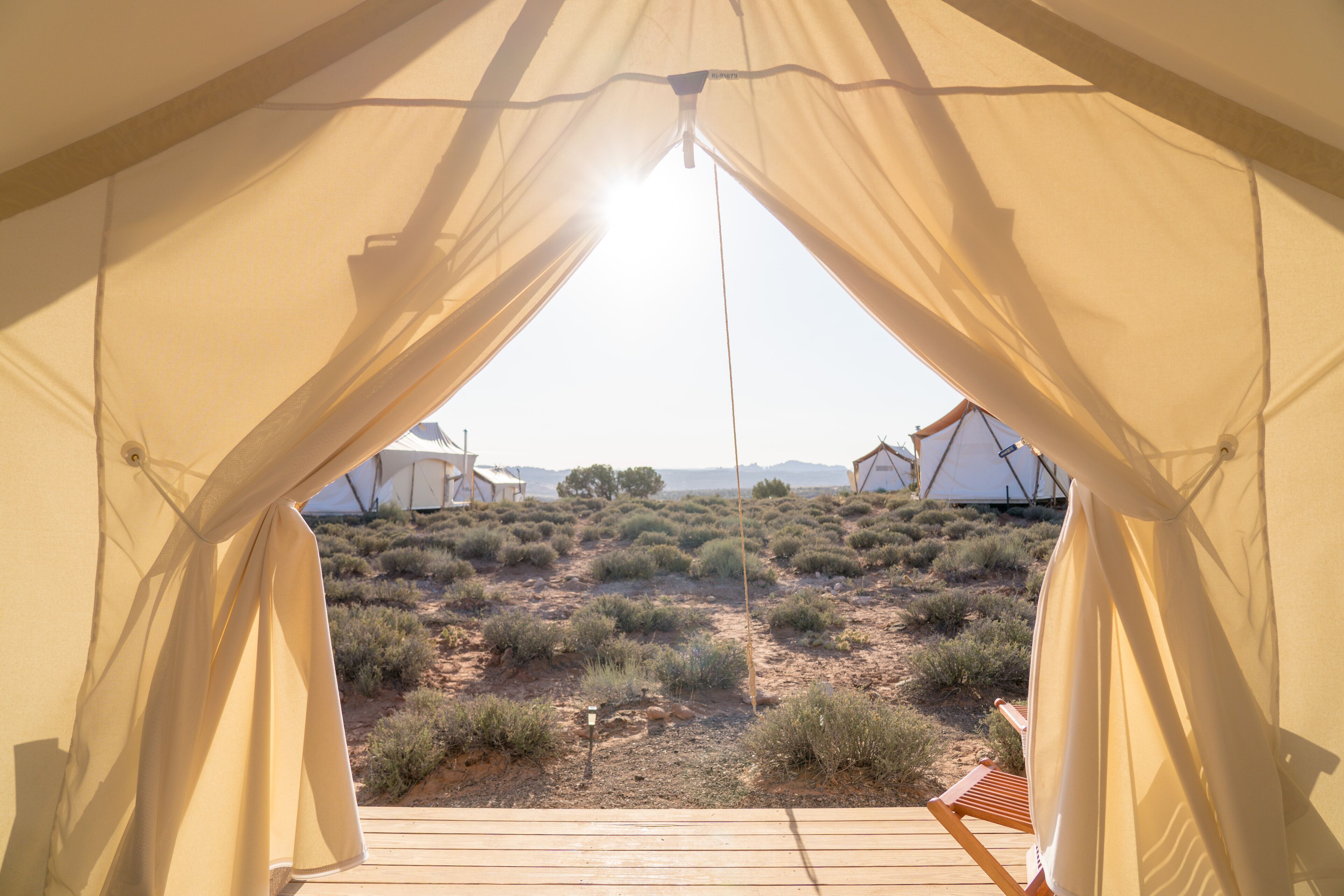 A tent with a wooden deck and beige curtains, overlooking a desert landscape with sparse vegetation and distant tents.