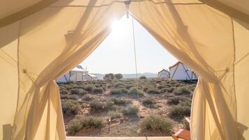 A tent with a wooden deck and beige curtains, overlooking a desert landscape with sparse vegetation and distant tents.