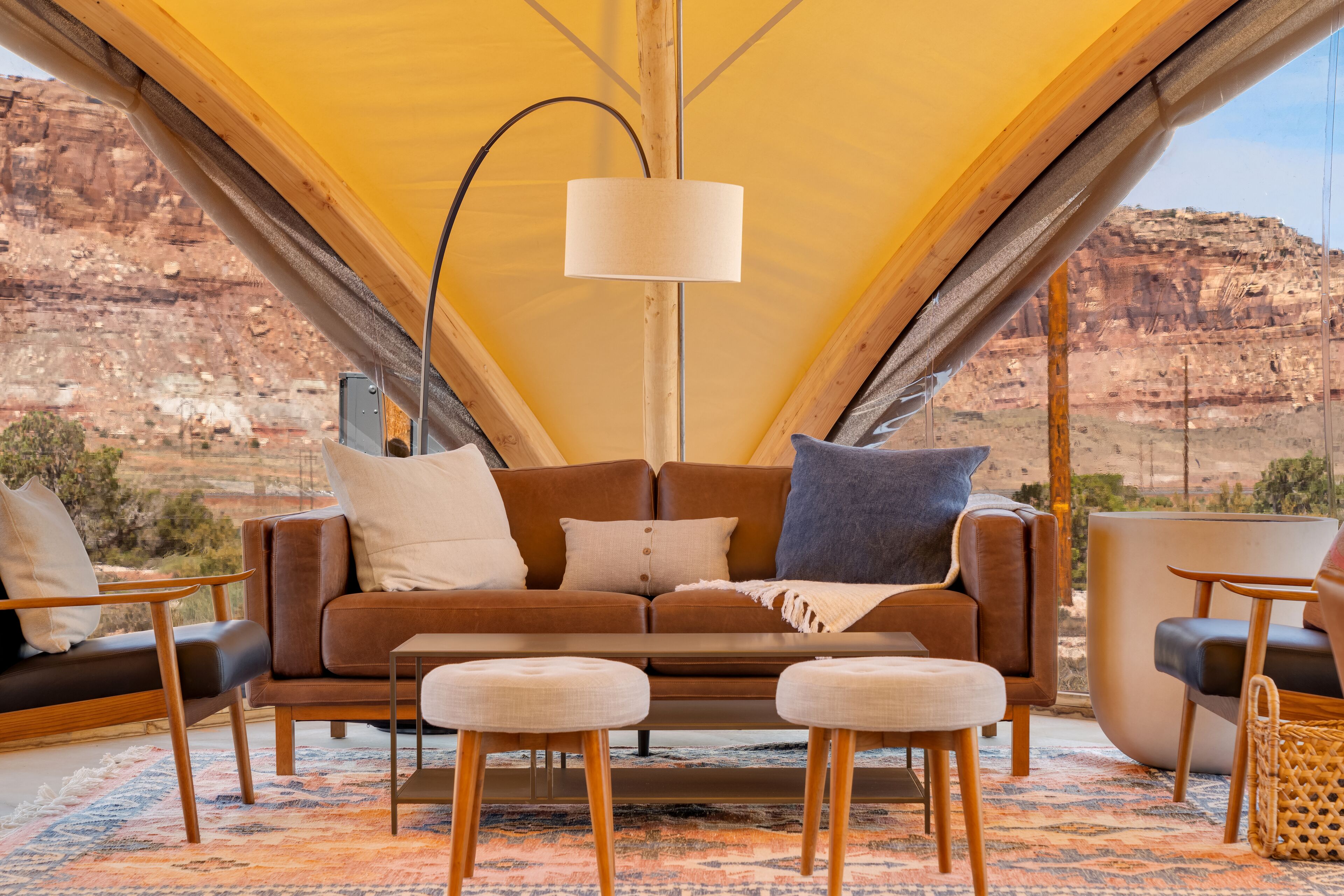 A tent interior with a brown sofa, two ottomans, a lamp, and a view of a rocky landscape.