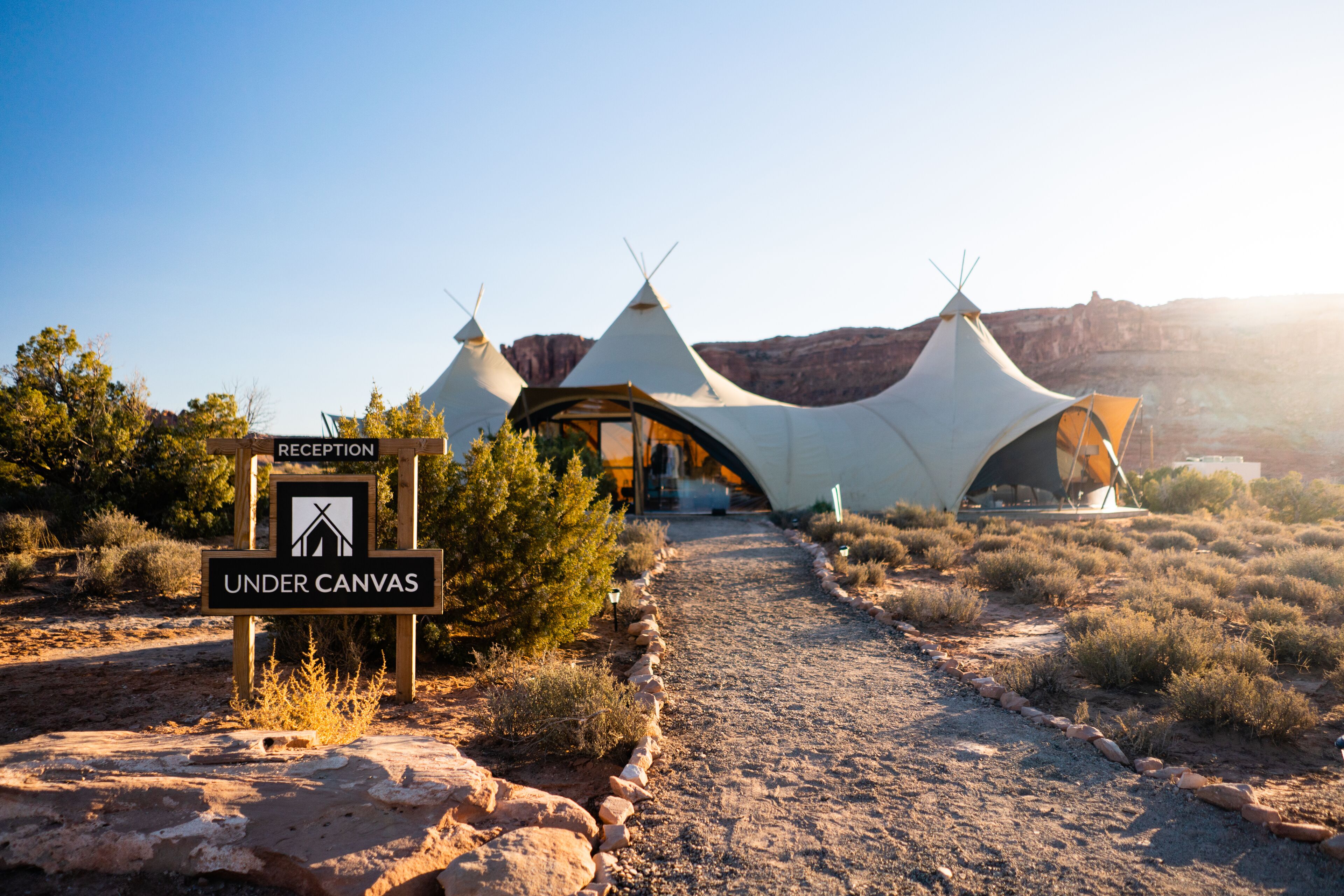 A reception sign and a sign for 'Under Canvas' in front of a series of white tents with pointed tops, set in a desert landscape.