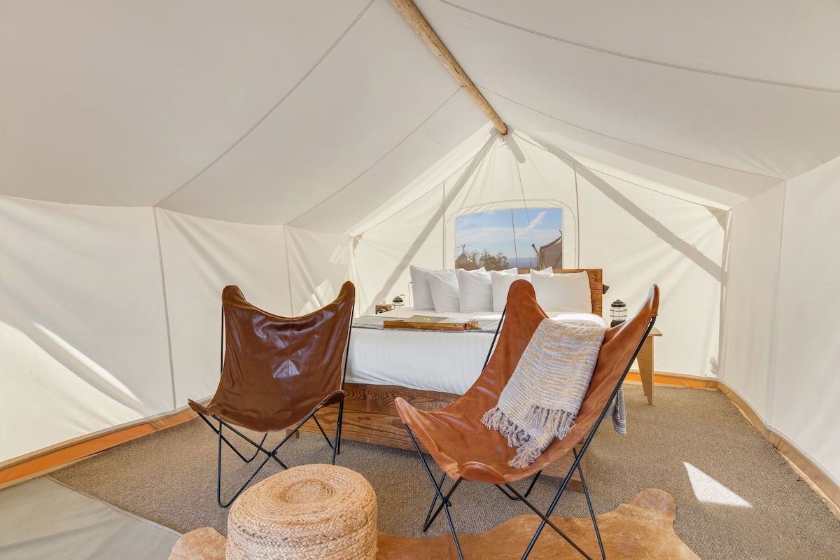 A tented room with a bed, two leather chairs, a woven ottoman, and a view of the landscape through the window.