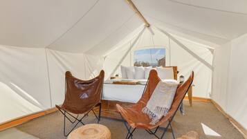 A tented room with a bed, two leather chairs, a woven ottoman, and a view of the landscape through the window.