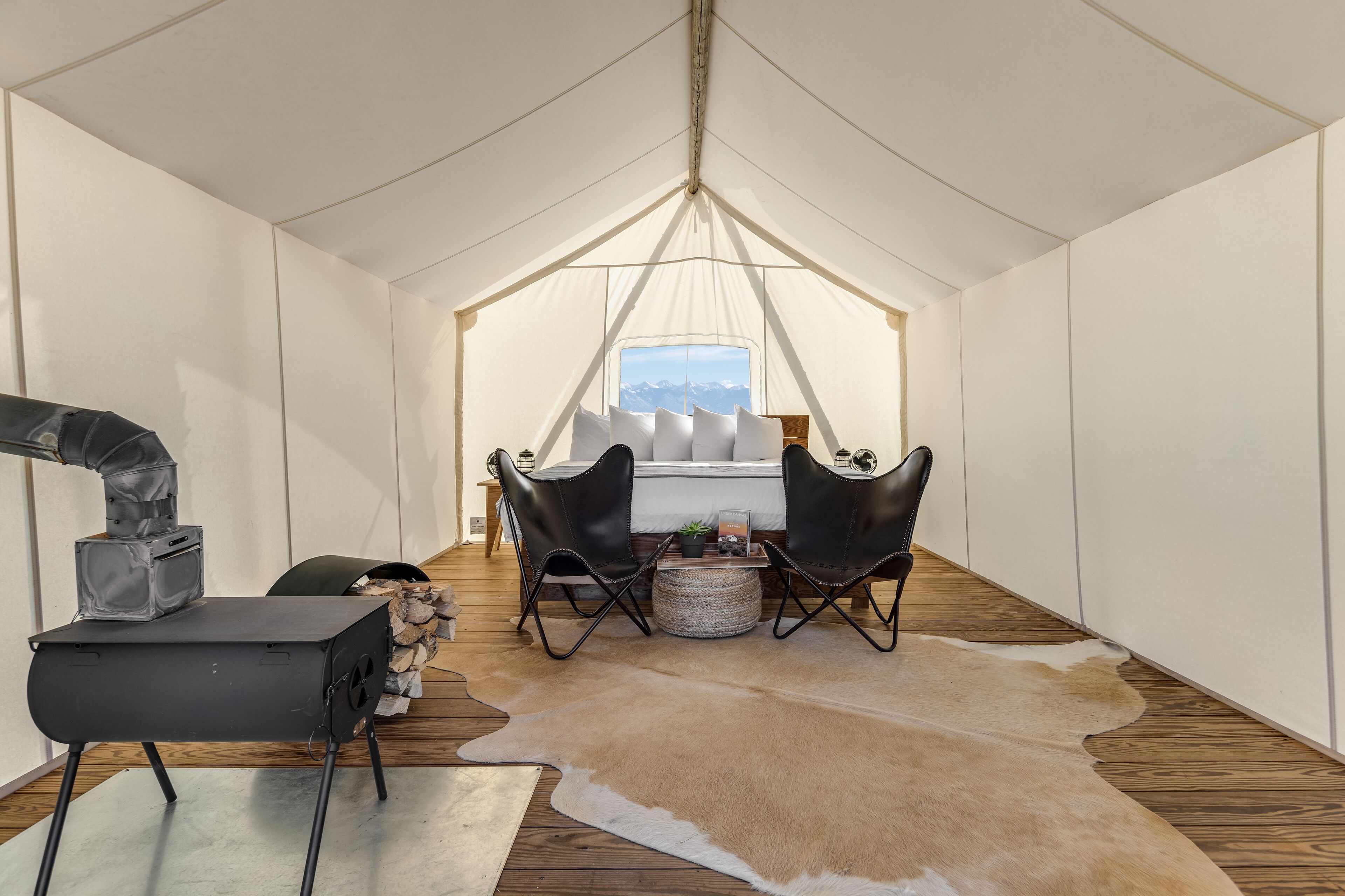 A tented room with a bed, two chairs, a heater, and a view of mountains.