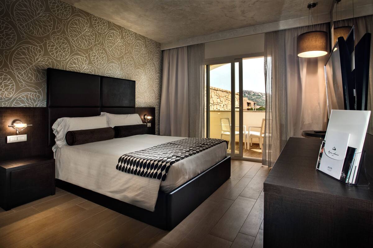 A modern hotel room with a large bed, a nightstand with a lamp, a desk with a computer, and a balcony with a view.