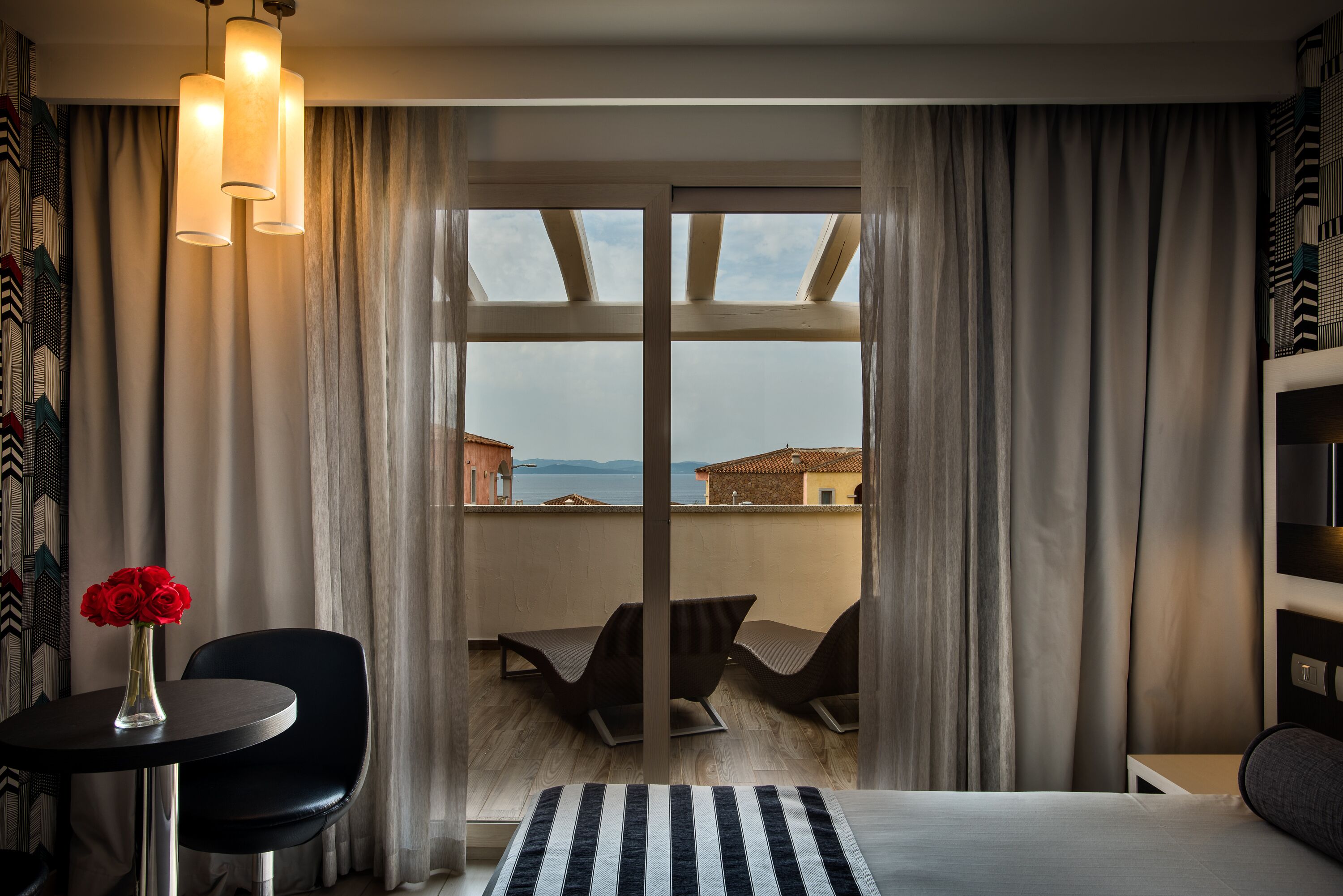 A hotel room with a bed, a small table with a vase of red roses, a chair, and a view of the sea through large windows.