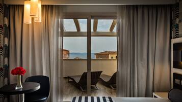 A hotel room with a bed, a small table with a vase of red roses, a chair, and a view of the sea through large windows.