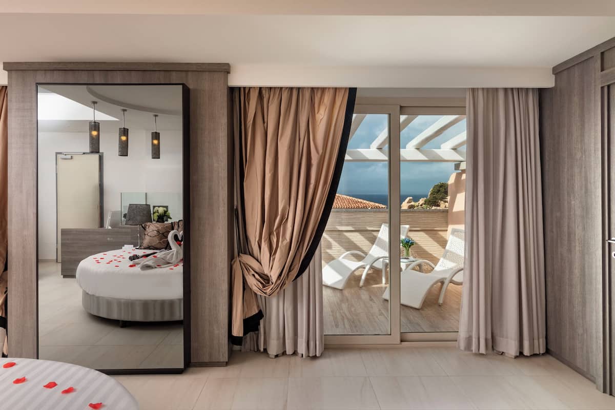 A modern hotel room with a large mirror, a bed with rose petals, a balcony with a view of the sea, and a dining area with white chairs.