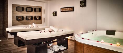 A spa room with two massage tables, a large bathtub with rose petals, and a 'comfort zone' storage unit.