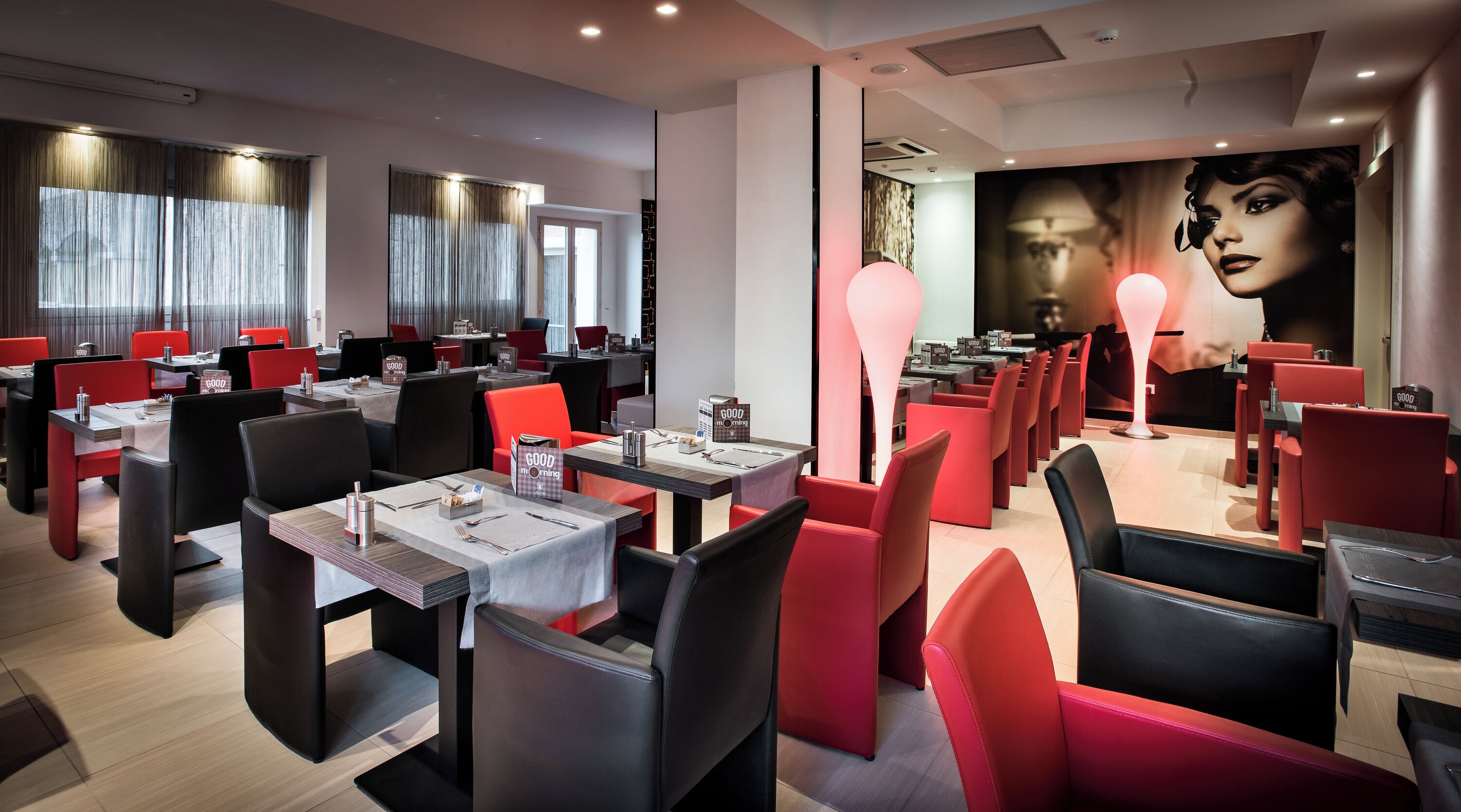 A modern restaurant interior with red and black chairs, tables with place settings, and a large mural on the wall.